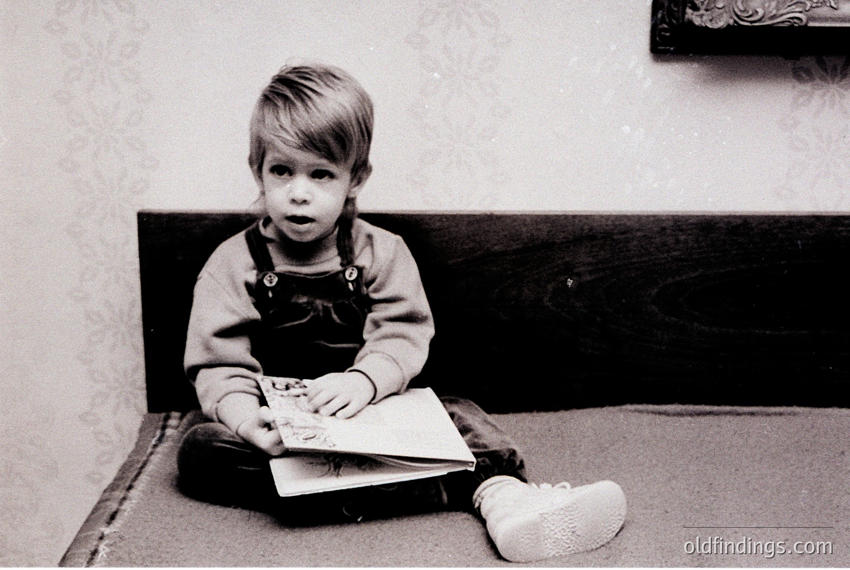 Black-and-white portrait of a young child (approx. 3-5 years) seated on a bed, holding an open book with a colorful illustration. Wearing overalls and white slip-on shoes, against a patterned wall backdrop. Likely mid-20th century (1950s–1970s) domestic setting.