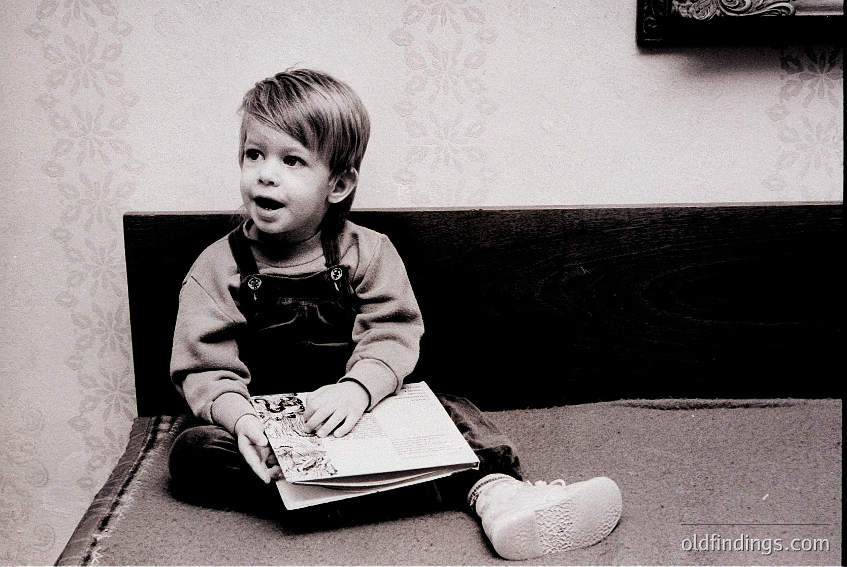 Black-and-white portrait of a young child (approx. 3-5 years) engrossed in a book, seated on a wooden bench. Overalls with buttons, white sneakers, and a neutral-toned sweater. Indoor setting with patterned wallpaper and framed artwork. Evokes mid-20th century domestic life.
