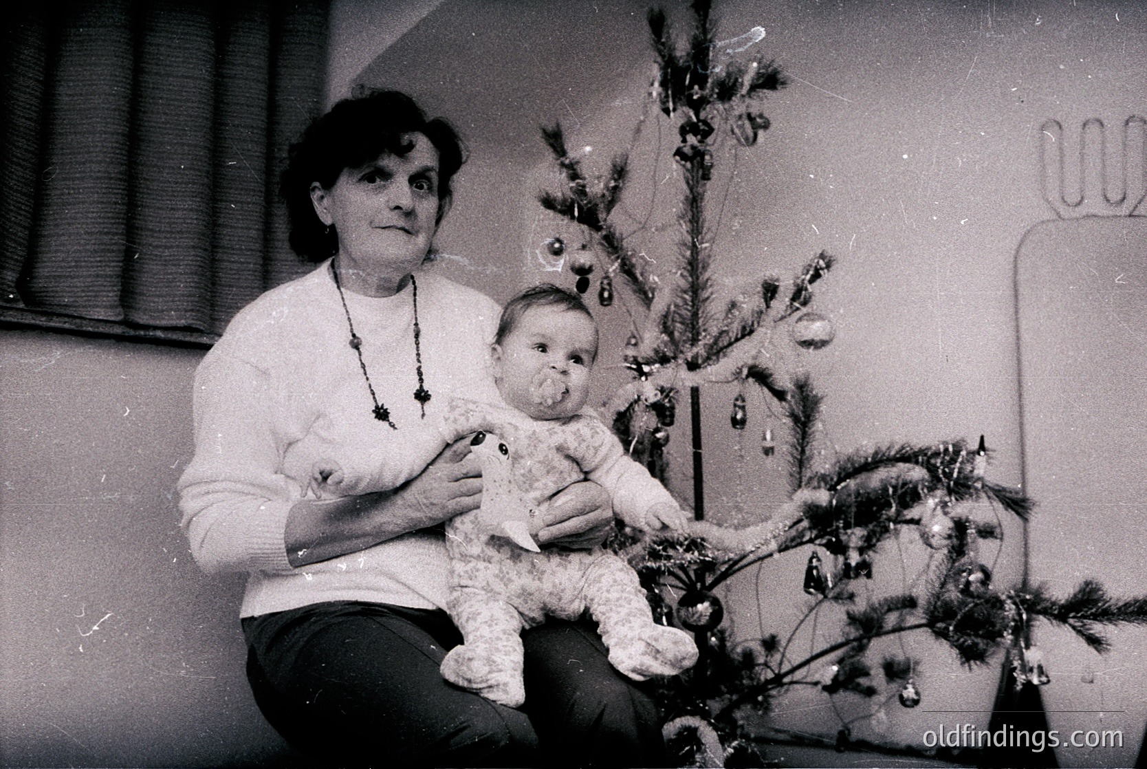 A woman cradles a baby beside a modestly decorated Christmas tree in an indoor setting, likely mid-20th century. The tree features simple ornaments and a single light bulb. The woman wears a necklace with circular pendants and a light sweater. [Vintage holiday portrait featuring a woman holding a baby beside a small Christmas tree with classic ornaments, mid-20th century ]