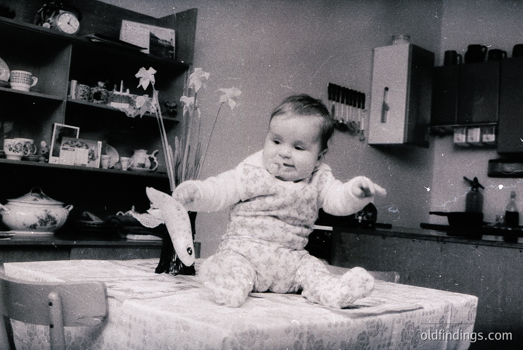Vintage black-and-white photo of a toddler in a floral onesie, seated on a wooden stool, holding a paper airplane. Surrounding objects include a shelf with decorative items, a clock, and a vase with dried flowers. Mid-20th century domestic interior.