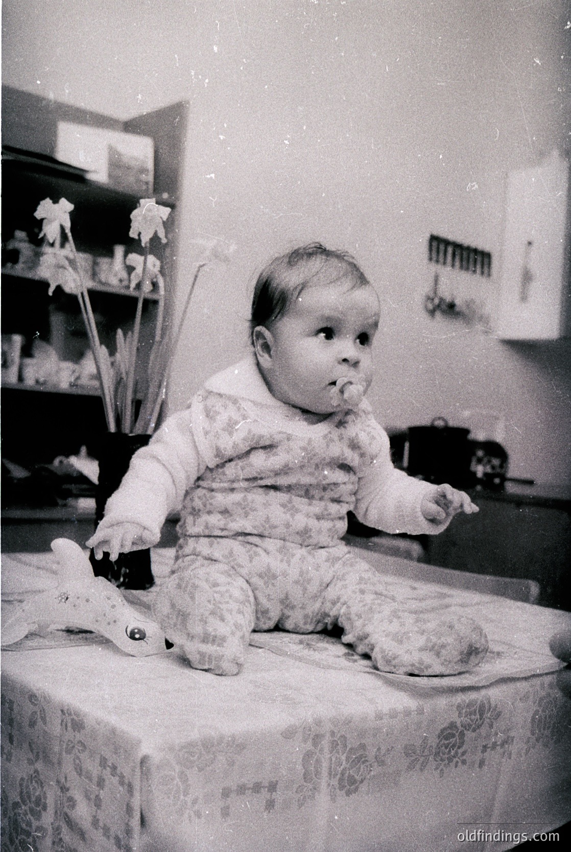 Vintage black-and-white photo of a toddler sitting on a patterned tablecloth, holding a toy. Indoor setting with floral wall decor and a shelf in background. Likely mid-20th century domestic scene.