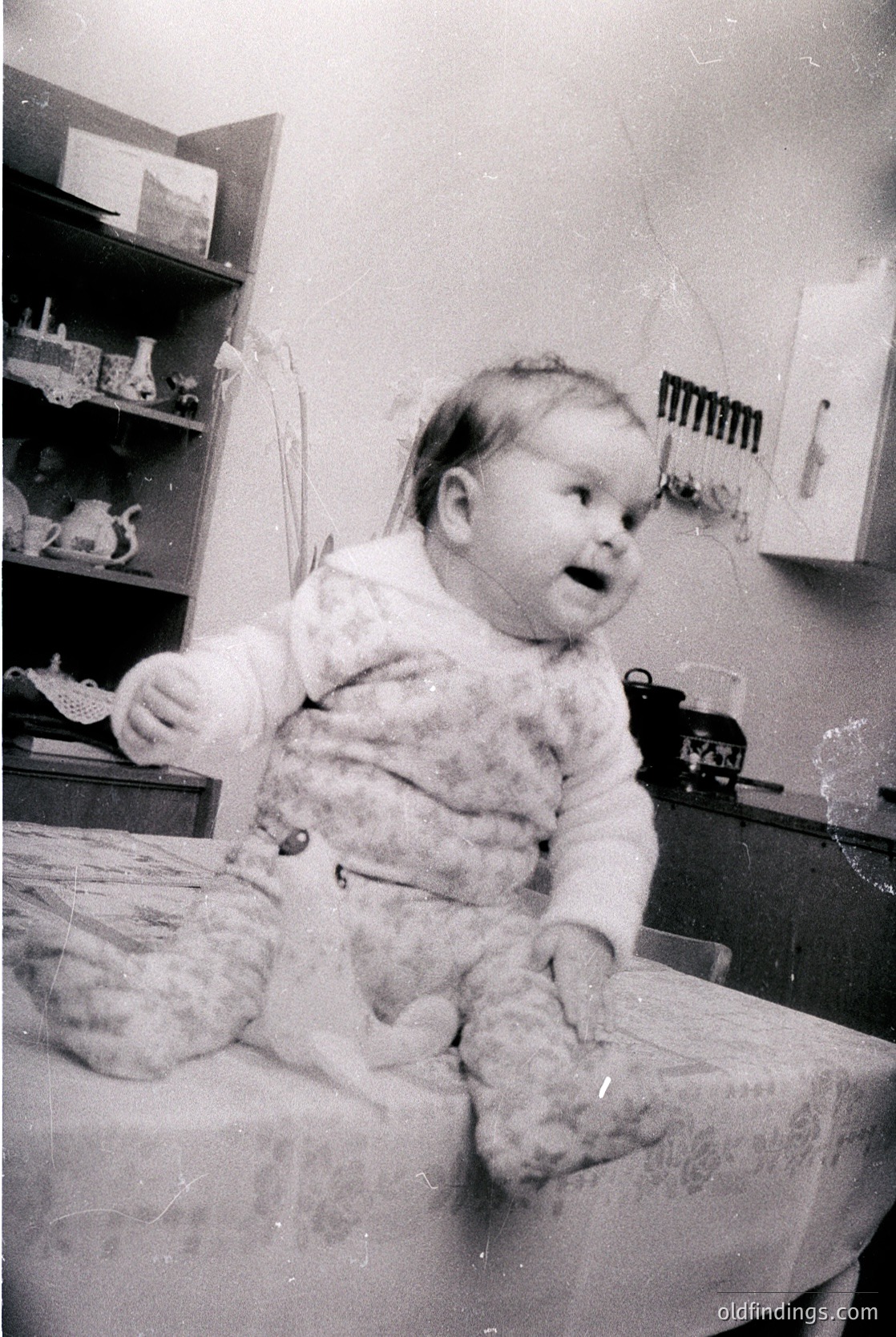 Vintage black-and-white photo of a toddler in a plush, textured sweater sitting on a highchair. Surrounding shelves display ceramic toys and kitchenware, suggesting a mid-20th-century domestic setting.