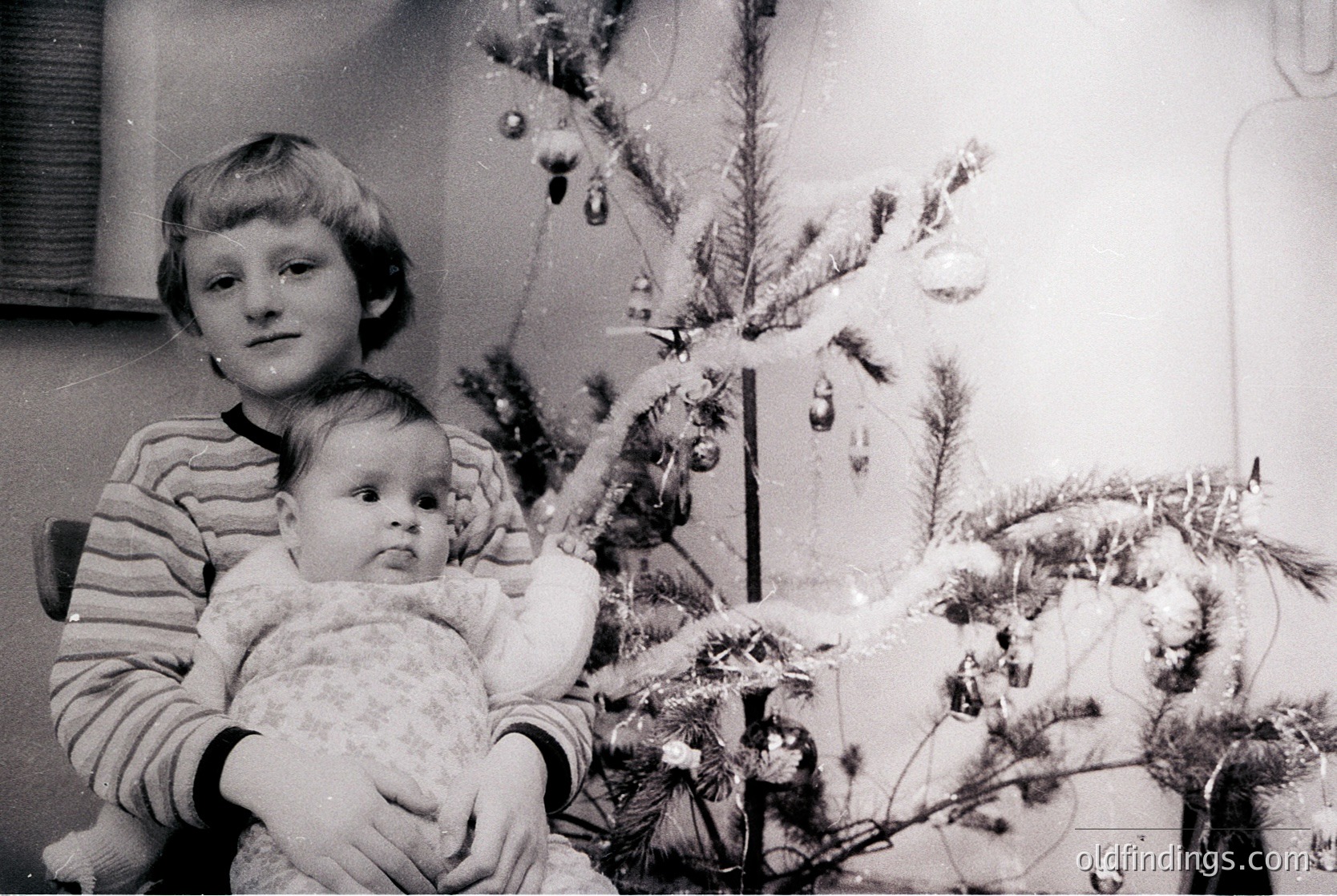 A mid-20th century (likely 1960s–1970s) black-and-white holiday scene: an older child holds a baby in front of a modestly decorated Christmas tree with tinsel and ornaments. Indoor setting suggests a cozy, family-centric atmosphere. Perfect for nostalgic or historical research on holiday traditions.