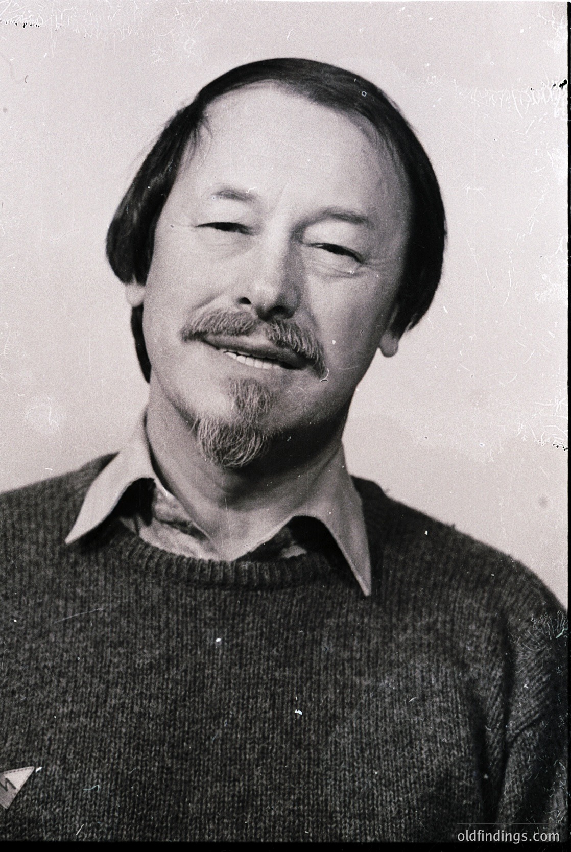 Portrait of a man with a relaxed expression, closed eyes, and a mustache/beard. Wearing a collared sweater with a subtle patch on the left lapel. Likely mid-20th century (1960s–1970s) based on hairstyle and attire.