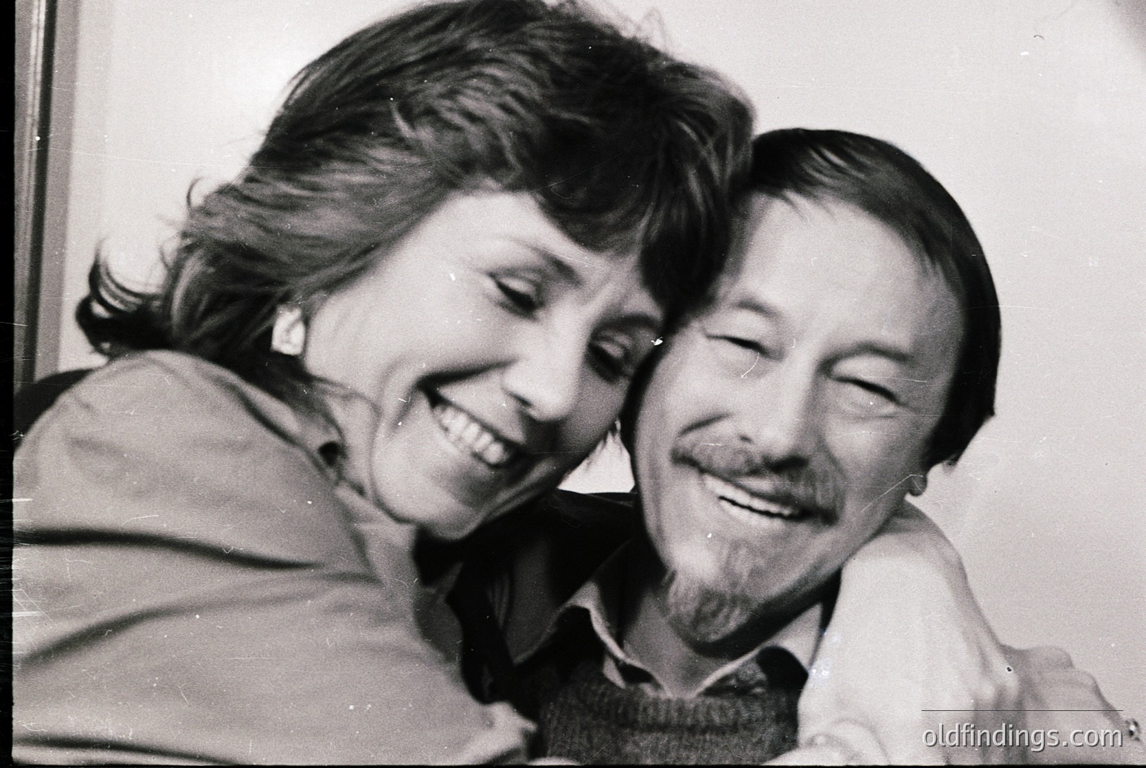 Black-and-white portrait of a joyful couple embracing indoors, likely mid-20th century. Woman’s short, voluminous hair and man’s mustache and sweater suggest 1960s–1970s fashion. Warm, candid expressions convey intimacy.