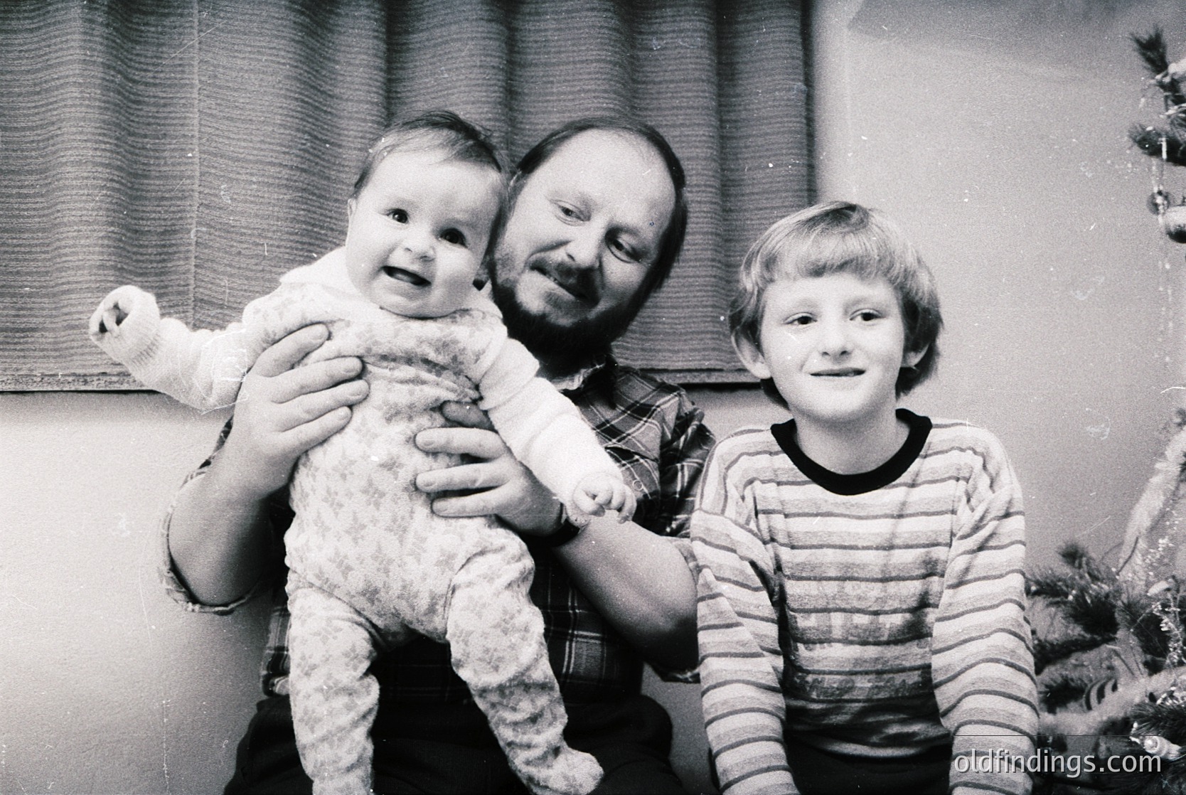 Family portrait from the 1970s–1980s, featuring a man holding a toddler while a young boy stands beside him. Indoor setting with holiday decorations (ornaments, curtain) suggesting winter. Casual attire: plaid shirt, striped sweater, and knitted outfit. Warm, candid moment capturing generational bonds.