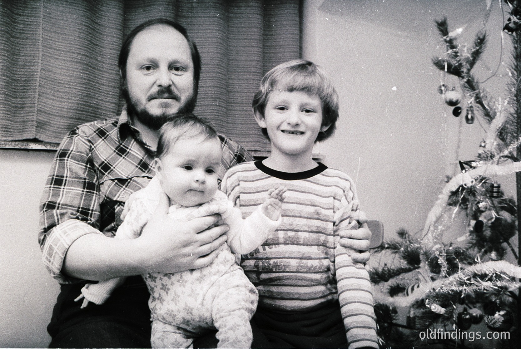 Family portrait from the 1970s–1980s featuring a man in a plaid shirt holding a baby, with a smiling boy in striped sweater beside them. Decorated Christmas tree with ornaments and tinsel in background. Indoor setting, likely residential.