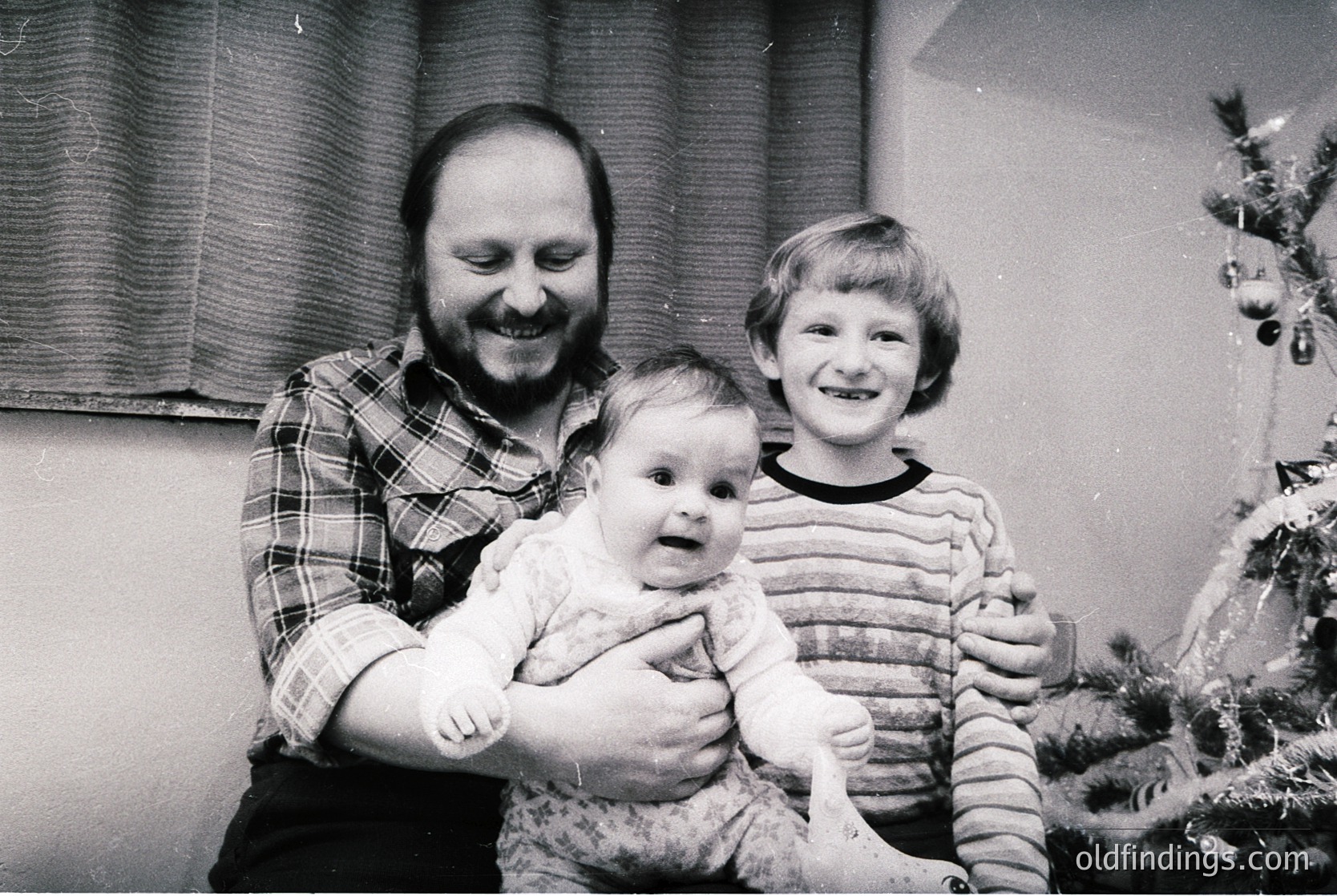 Black-and-white family portrait featuring a man in a plaid shirt holding a baby, with a smiling child beside him. Decorated Christmas tree and festive curtains in background. Likely 1970s–1980s holiday scene.