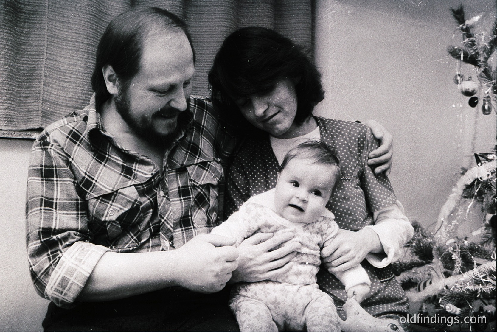 Family portrait featuring a man in a plaid shirt, woman in a patterned blouse, and a baby in a knitted outfit, posed indoors beside a decorated Christmas tree. Mid-20th century holiday setting, likely 1960s–1970s.