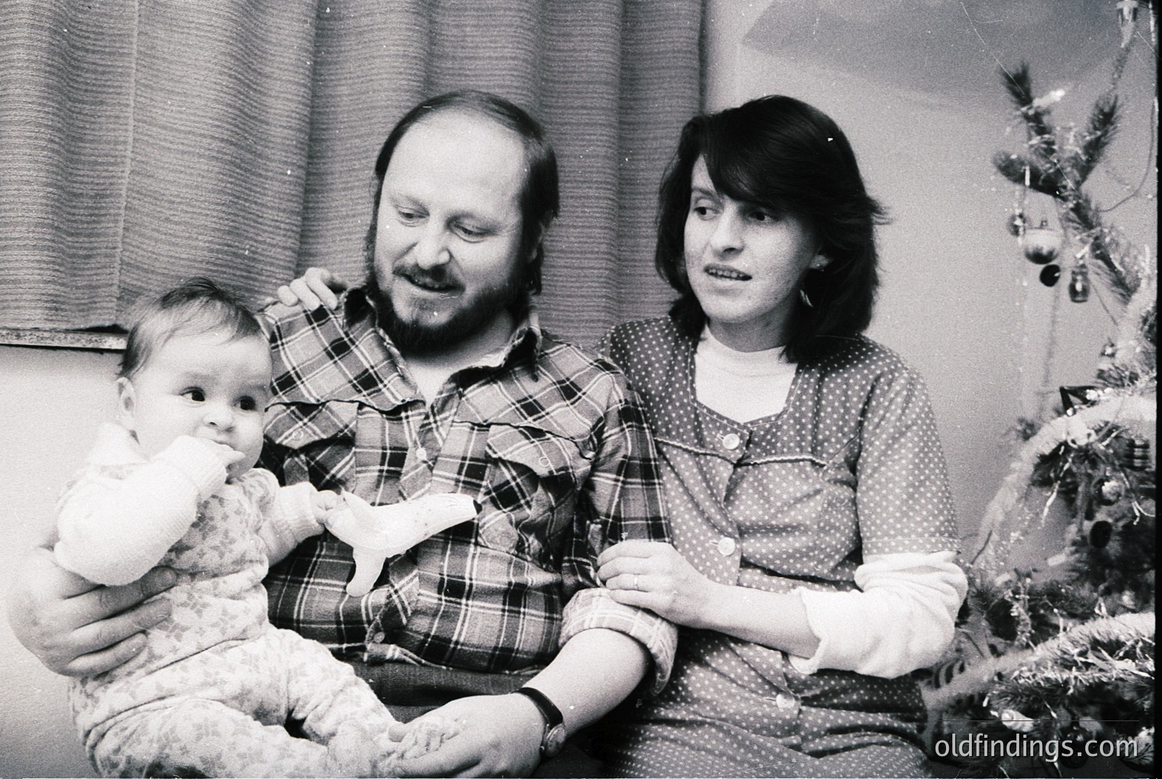 A black-and-white family portrait from the **1970s–1980s**, featuring a man in a plaid shirt, a woman in a patterned blouse, and a baby in a white outfit. They pose indoors near a decorated Christmas tree with ornaments. Warm, candid holiday atmosphere.