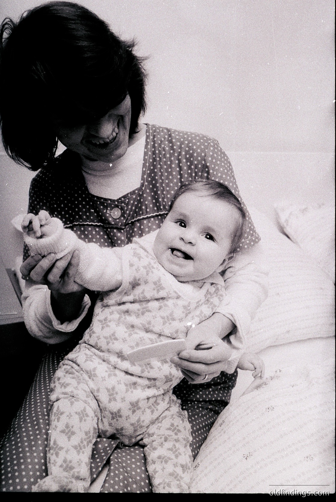 A tender black-and-white moment: a woman in a polka-dotted dress cradles an infant in floral pajamas, both smiling. Mid-20th century (1950s–1960s) domestic intimacy captured.