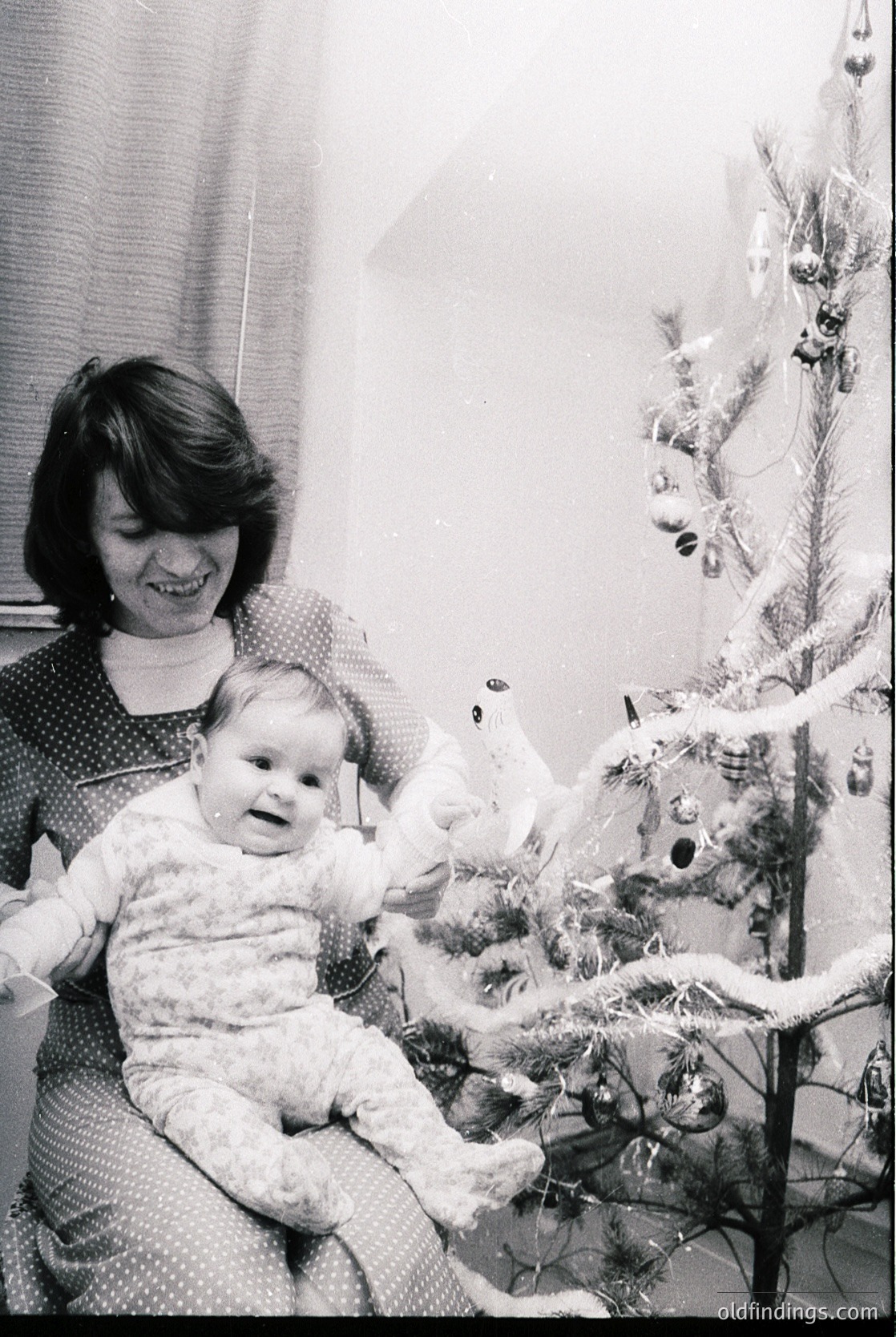 A woman in a patterned dress cradles a baby in a vintage holiday setting. Ornate Christmas tree with glass baubles and tinsel. Indoor, likely mid-20th century home decor.