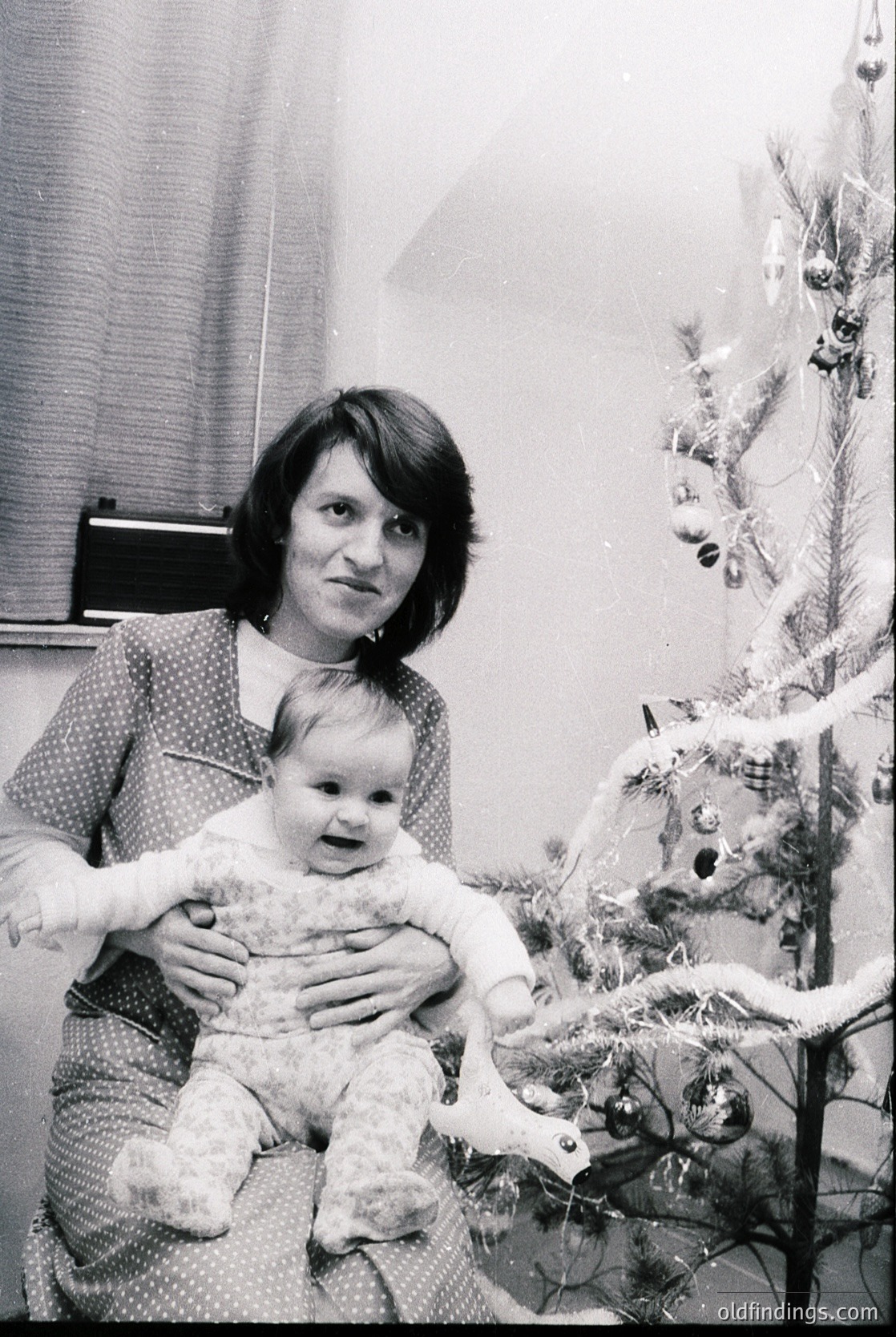 A woman in a patterned dress holds a baby in a festive indoor setting, likely during the 1970s. A decorated Christmas tree with ornaments and tinsel fills the background. The scene captures a timeless holiday moment.