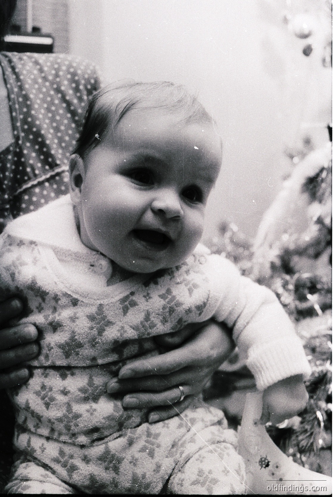 Black-and-white portrait of an infant in mid-1960s attire, wearing a patterned long-sleeve bodysuit and knitted sweater. The child’s expression suggests surprise or excitement. Background shows blurred floral wallpaper and partial adult figure in a polka-dot dress.
