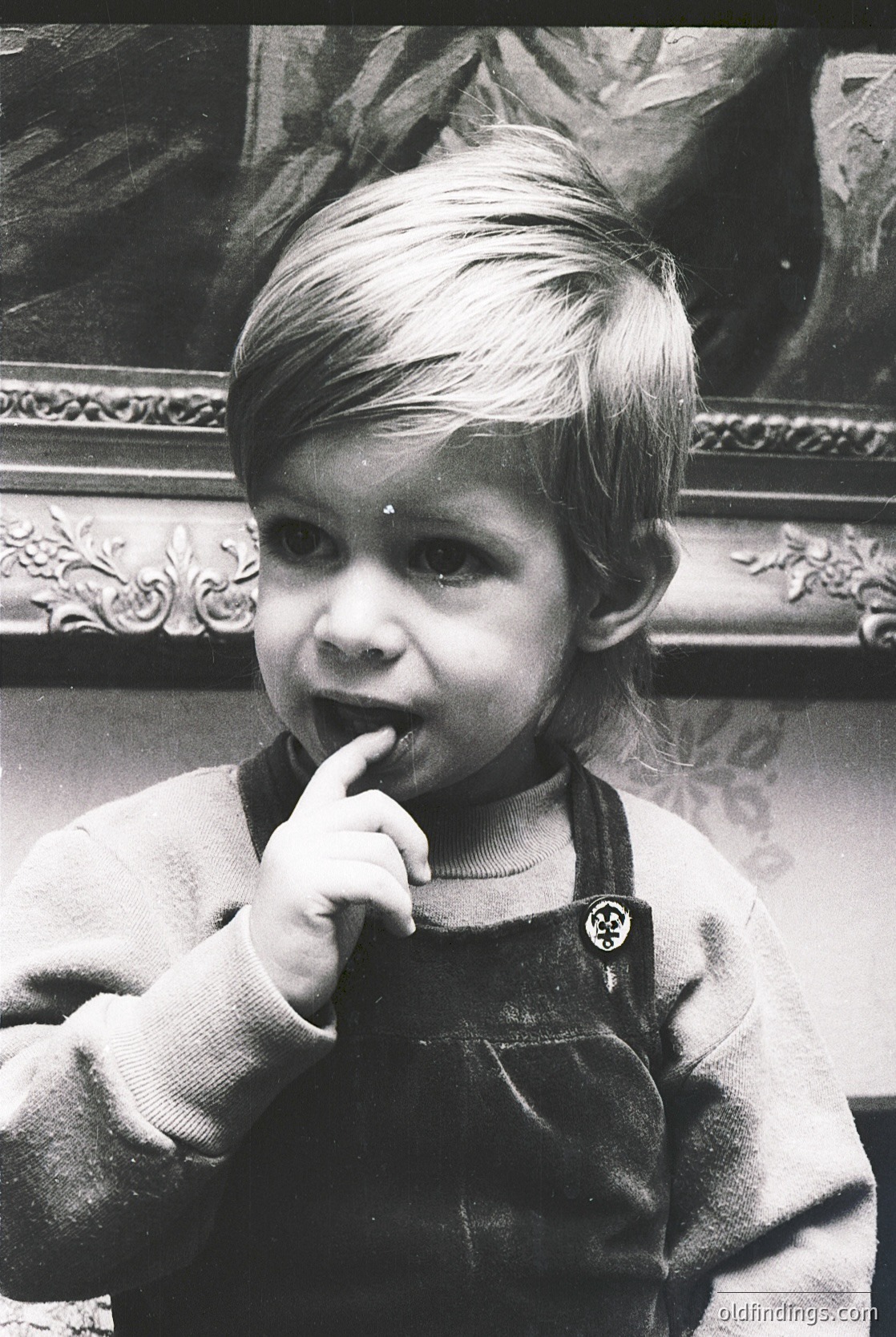 Young child in vintage overalls, mid-20th century style, posing indoors with ornate gilded frame background. Candid expression suggests curiosity or contemplation. Classic black-and-white portrait evokes mid-century family photography.