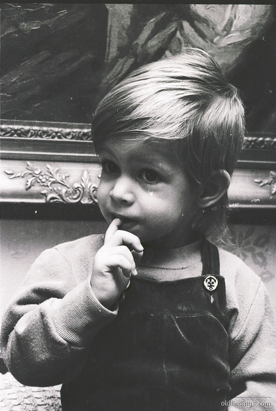 Vintage black-and-white portrait of a young child in a simple apron, posing with a finger in mouth. Ornate gilded frame and classical mural in background suggest mid-20th century European interior. Candid, nostalgic expression captures innocence and timelessness.