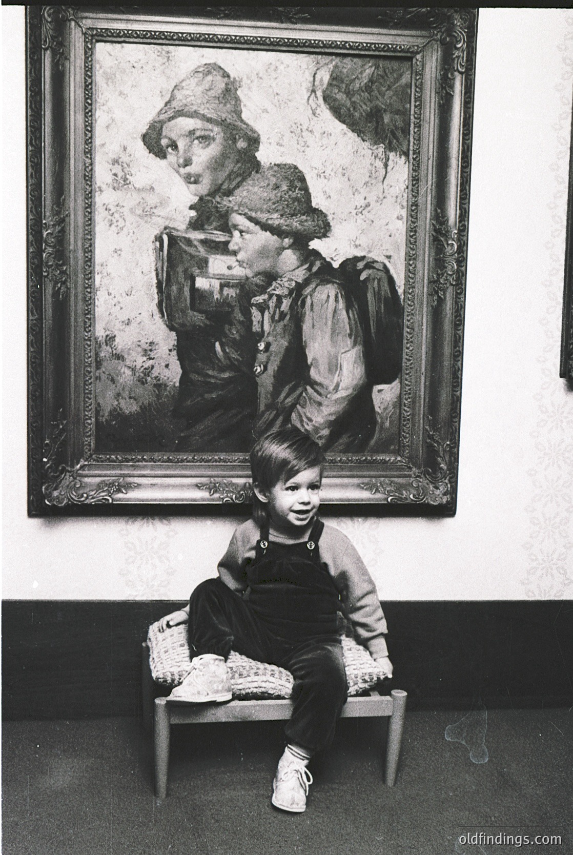 Vintage portrait of a child seated on a simple wooden stool, posing beside a framed painting of a woman in period attire (1950s-60s). The child wears overalls, a sweater, and white shoes, while the painting’s subject holds a camera and wears a wide-brimmed hat. Indoor setting with neutral walls, likely a studio or gallery.