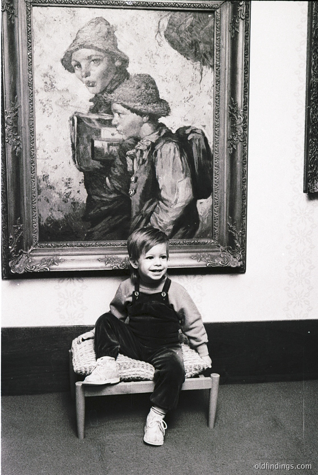 Vintage portrait of a young boy seated on a stool before a framed painting of a woman in outdoor attire, likely 1950s–1960s. The boy wears overalls and sneakers, while the painting depicts a woman in a fur hat and vintage coat. Indoor setting with neutral tones.
