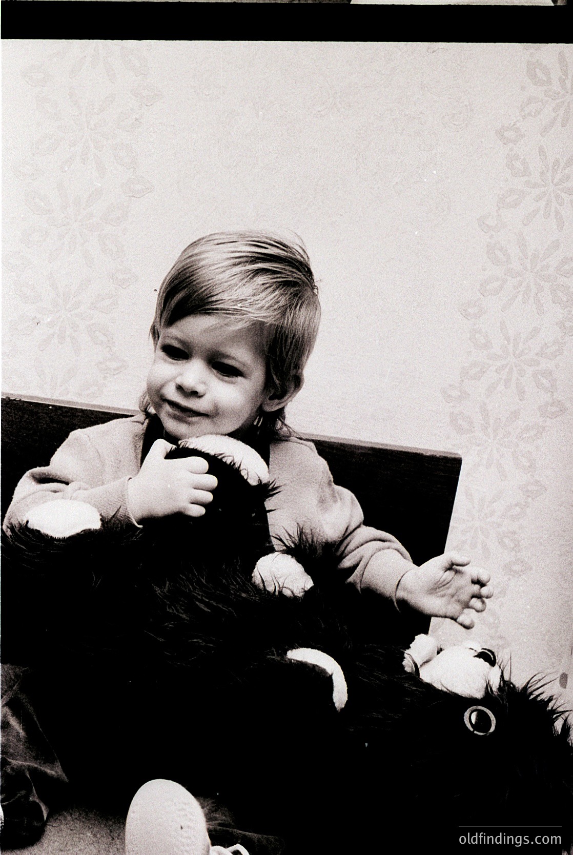 Vintage black-and-white photo of a toddler (approx. 2-3 years) sitting on a patterned sofa, clutching a plush toy. Wallpaper features floral motifs. Likely mid-20th century domestic setting.