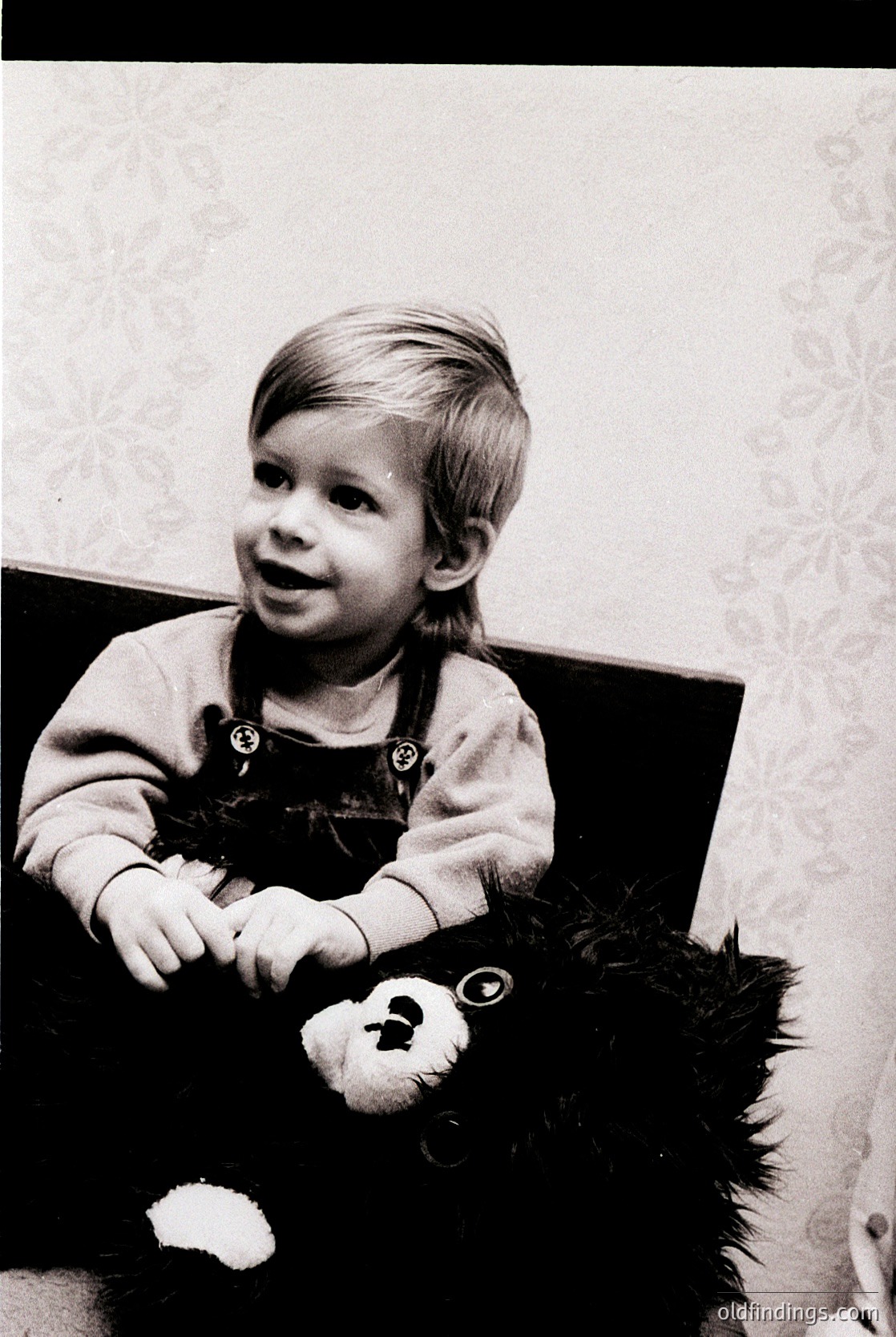Young child in 1970s-style overalls holding a vintage teddy bear with fur trim, seated indoors against patterned wallpaper. Candid, nostalgic family portrait.
