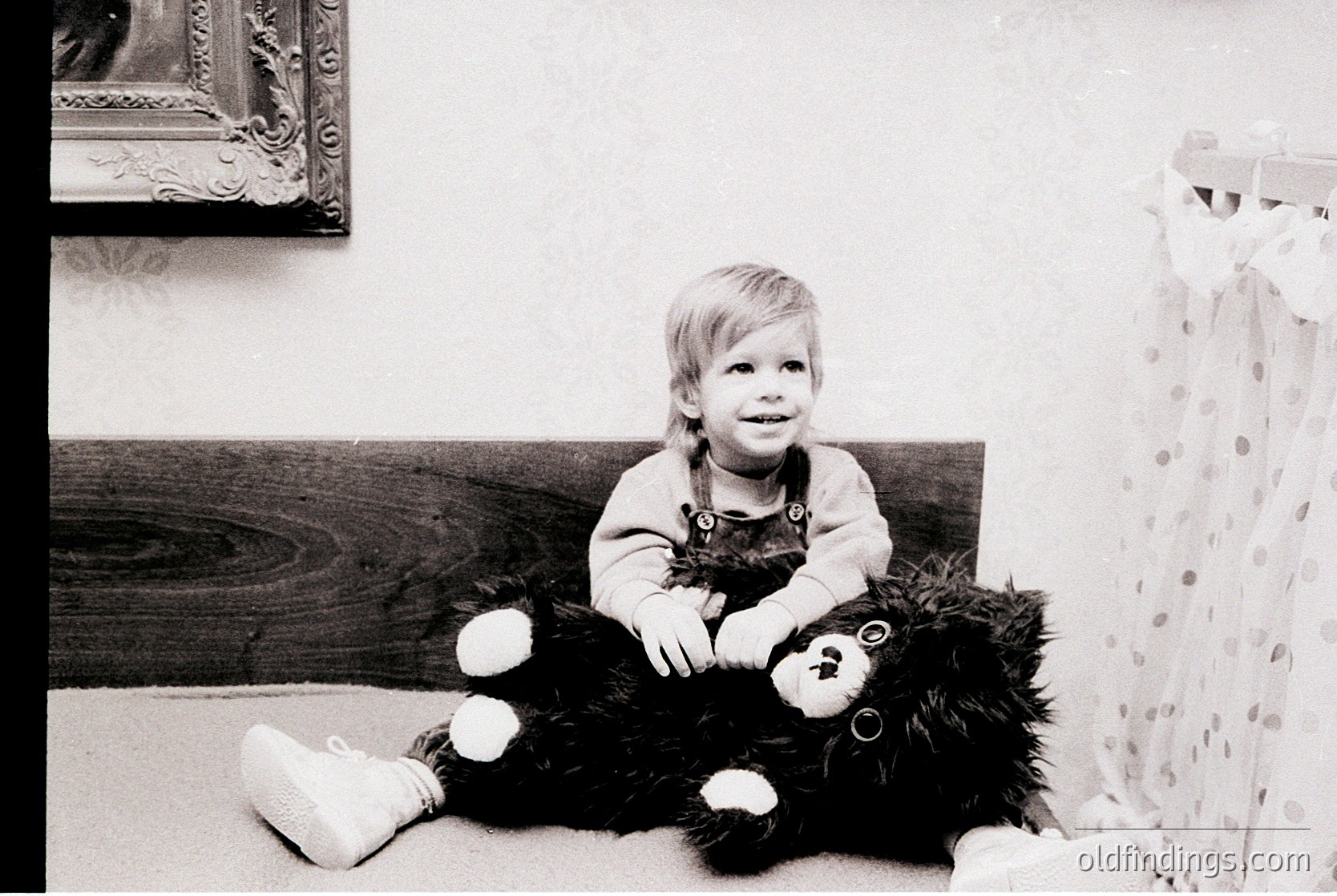 Vintage black-and-white portrait of a young child hugging a large teddy bear in a cozy indoor setting. Wooden headboard and floral-patterned bedding suggest a mid-20th-century home. Warm, candid expression captures innocence and playfulness.