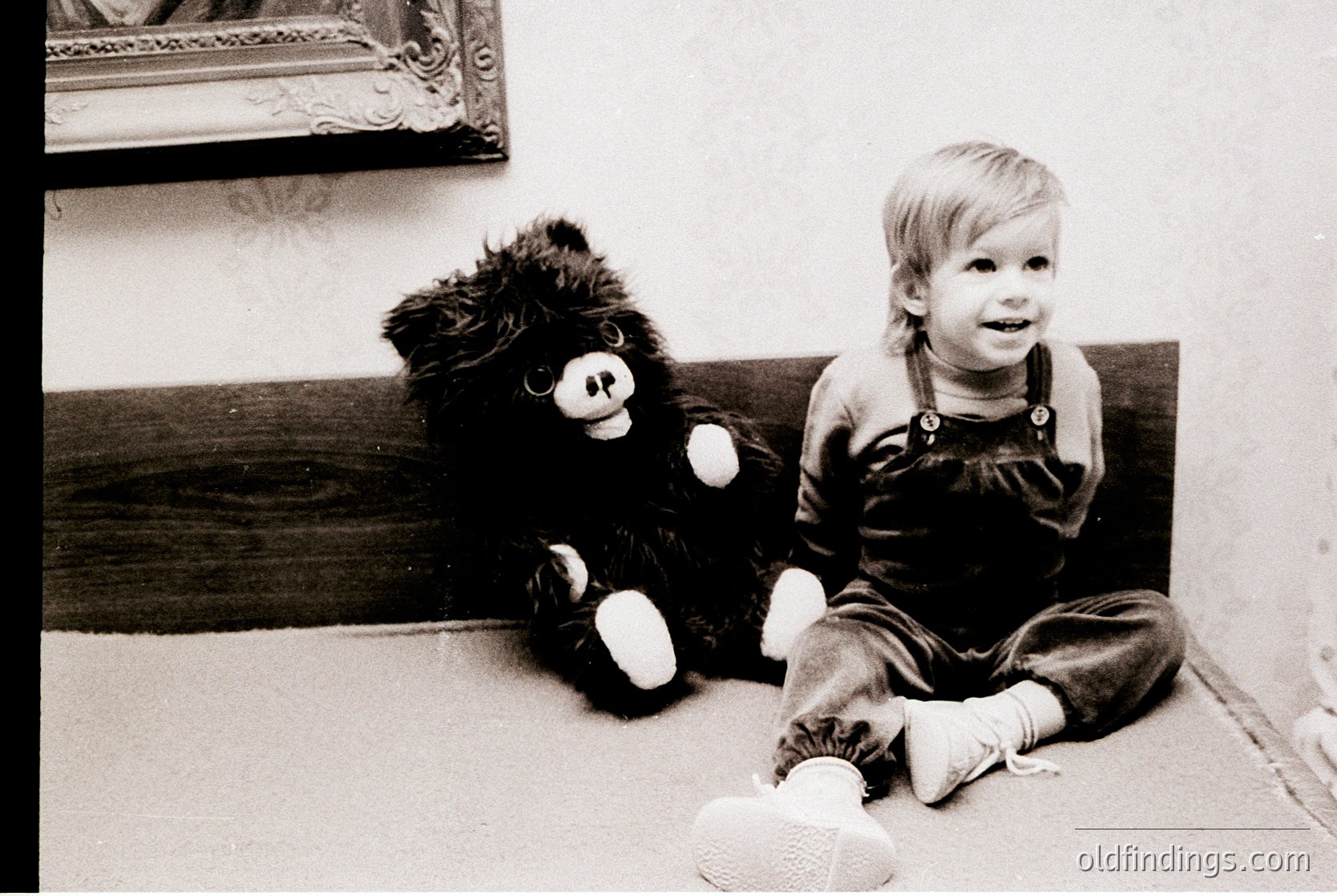 Black-and-white portrait of a young child (approx. 2-3 years) sitting beside a vintage stuffed bear with white paws and face patches. Indoor setting with wooden furniture and framed artwork in background. Likely mid-20th century (1950s–1970s) based on clothing and toy style.