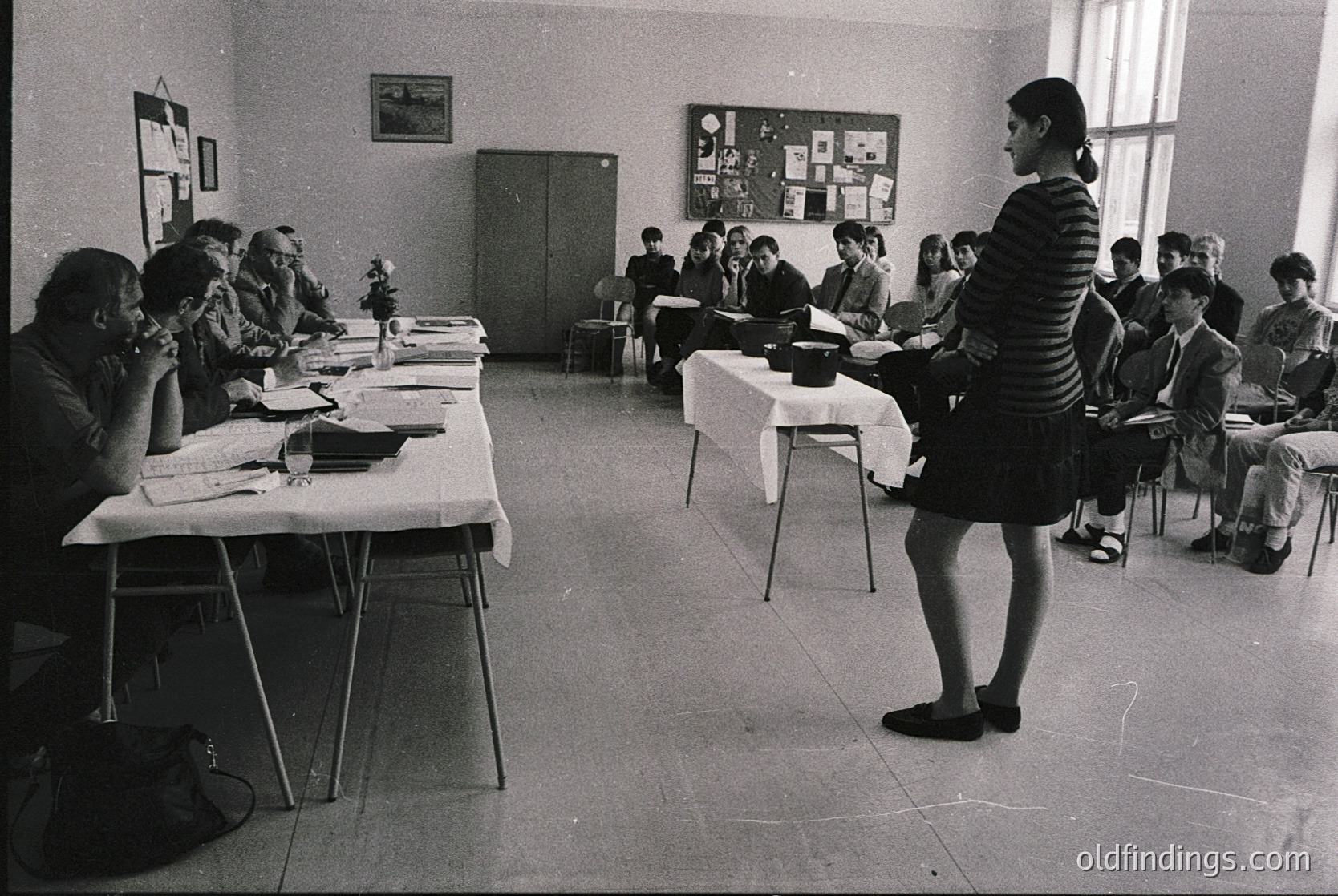 Classroom setting with students seated at long tables, engaged in a lecture or discussion. A teacher in a striped dress stands at a table, gesturing. Mid-20th century (likely 1960s–1970s) institutional or educational environment. Bulletin boards with posters and maps adorn walls.