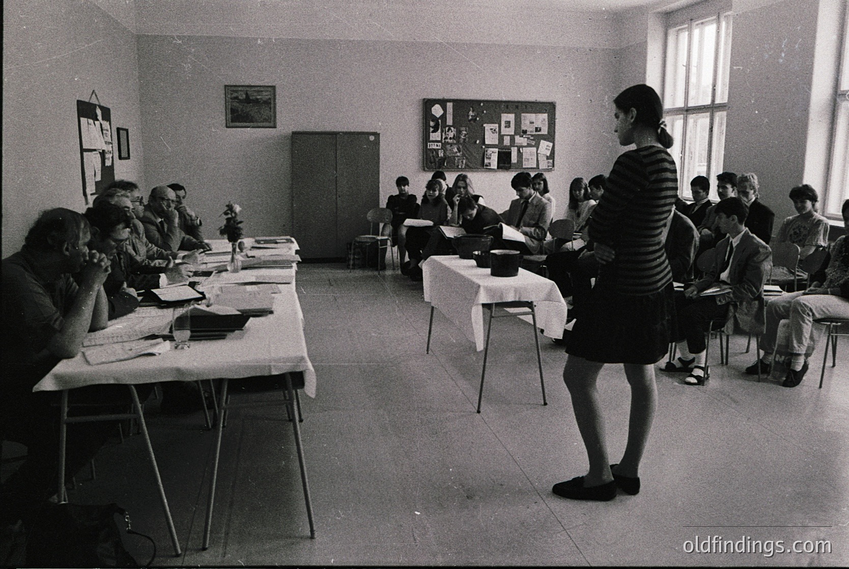 Classroom setting with mid-20th century institutional furniture, likely 1960s–1970s. Teacher in striped dress addresses seated students at long tables, surrounded by framed posters. Plain walls, fluorescent lighting, and uniform desks suggest a public or vocational school.