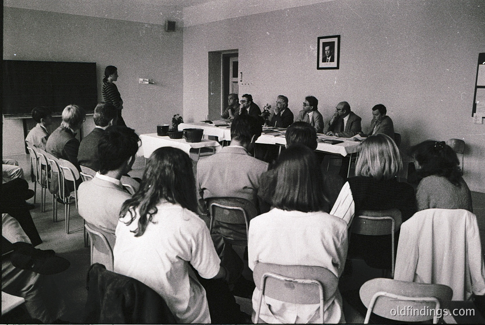Formal conference or meeting in a mid-century institutional setting, likely 1960s–1970s. Group of seated attendees facing a panel of speakers at a long table, framed by plain walls and a framed portrait. Attire suggests professional or academic context.