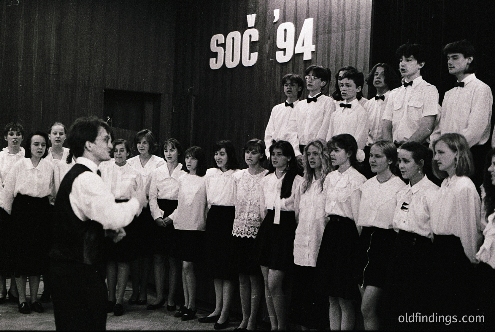 Young choir in formal attire performs on stage, 1994 Soči event. Uniforms feature white blouses, bow ties, and dark vests. Stage backdrop displays "Soči '94" in bold letters.