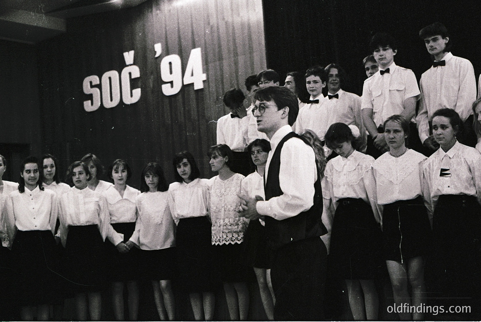 Young choir performing on stage, 1970s Czechoslovakia. Uniformed group in formal attire—men in vests, women in blouses and skirts—under "SČS '94" banner. Indoor venue with curtain backdrop. ČS