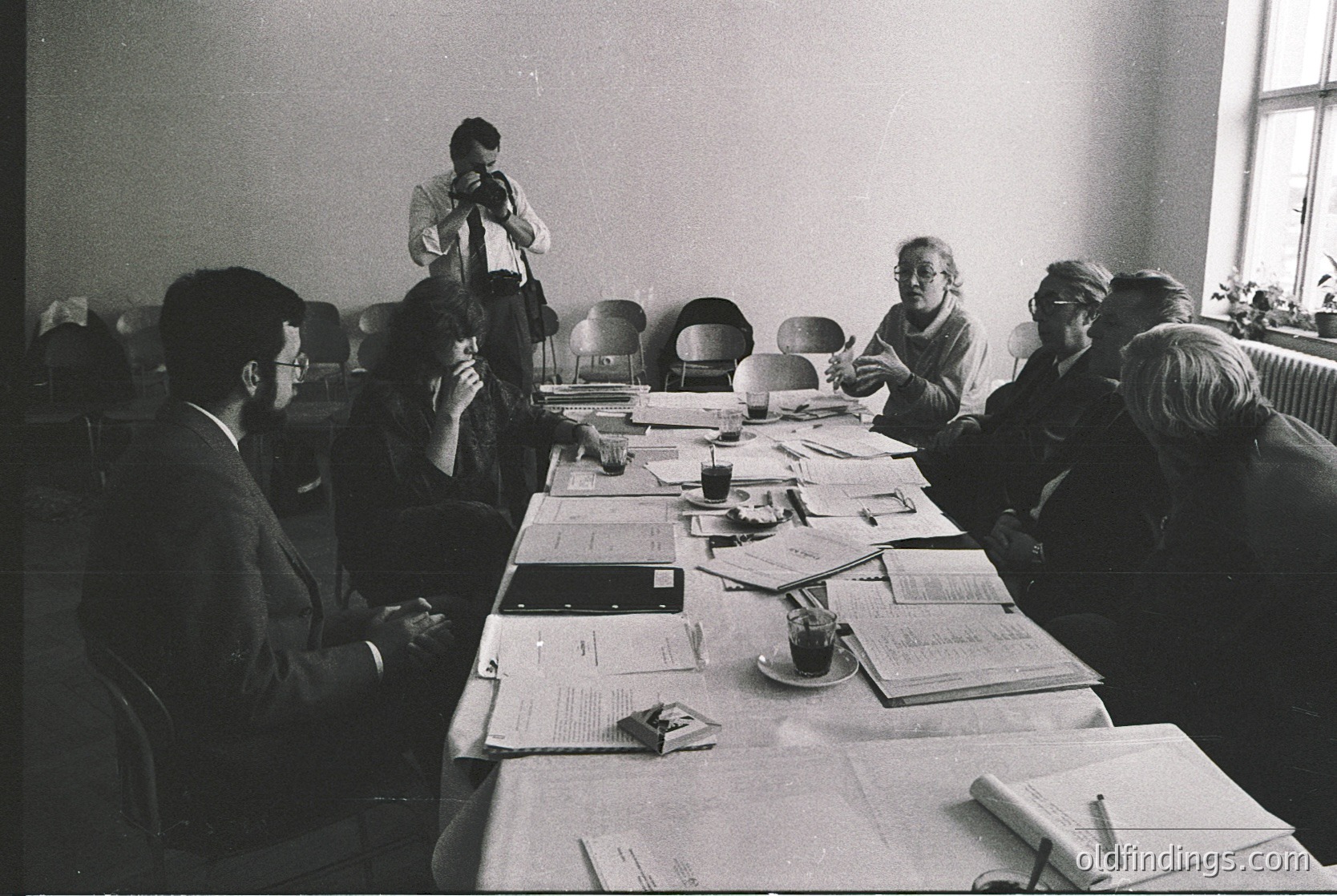 Mid-20th century office meeting with six professionals seated around a long table covered in documents. One man stands, speaking into a phone. Formal attire—suits, ties, and glasses—suggests a corporate or governmental setting. Typewriters and coffee cups indicate pre-digital workflows. Likely Eastern Bloc or Cold War-era office, 1960s–1980s.