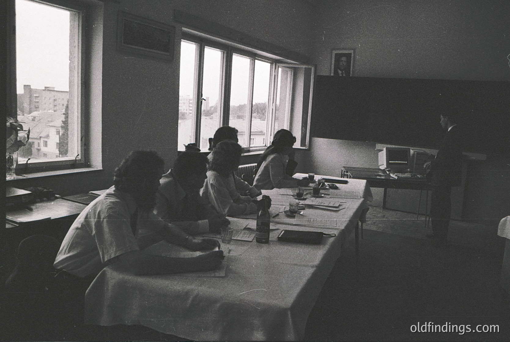Vintage black-and-white classroom scene with five seated students focused on individual workstations. Long table with typewriters, papers, and small bottles. Teacher stands at front, gesturing. Large windows reveal urban rooftops. Mid-century institutional design. Likely Eastern Bloc education setting, 1960s–1970s.