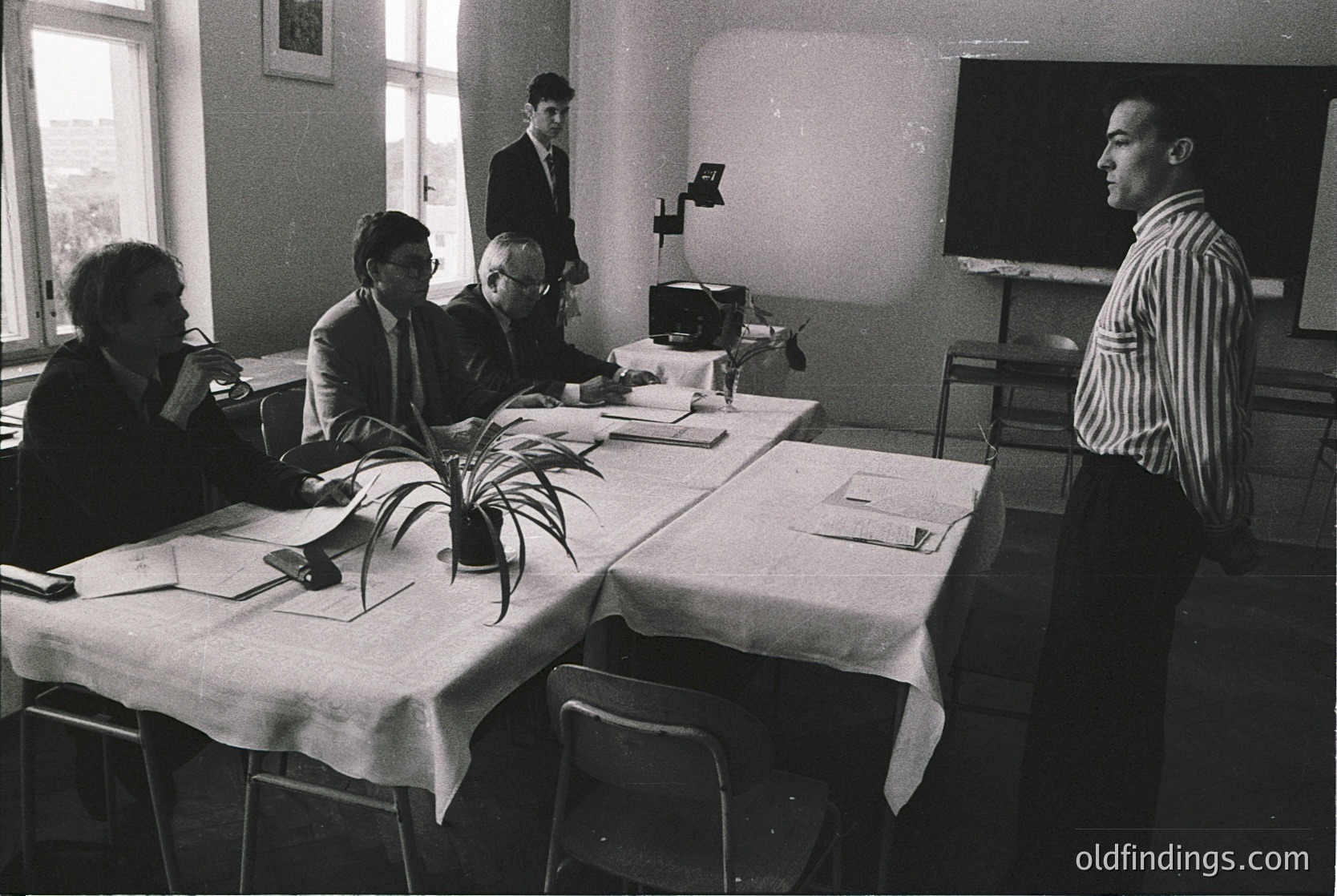 Mid-20th century conference room with four men in formal attire (1960s-1970s). White tablecloths, vintage projector, and classic office decor.