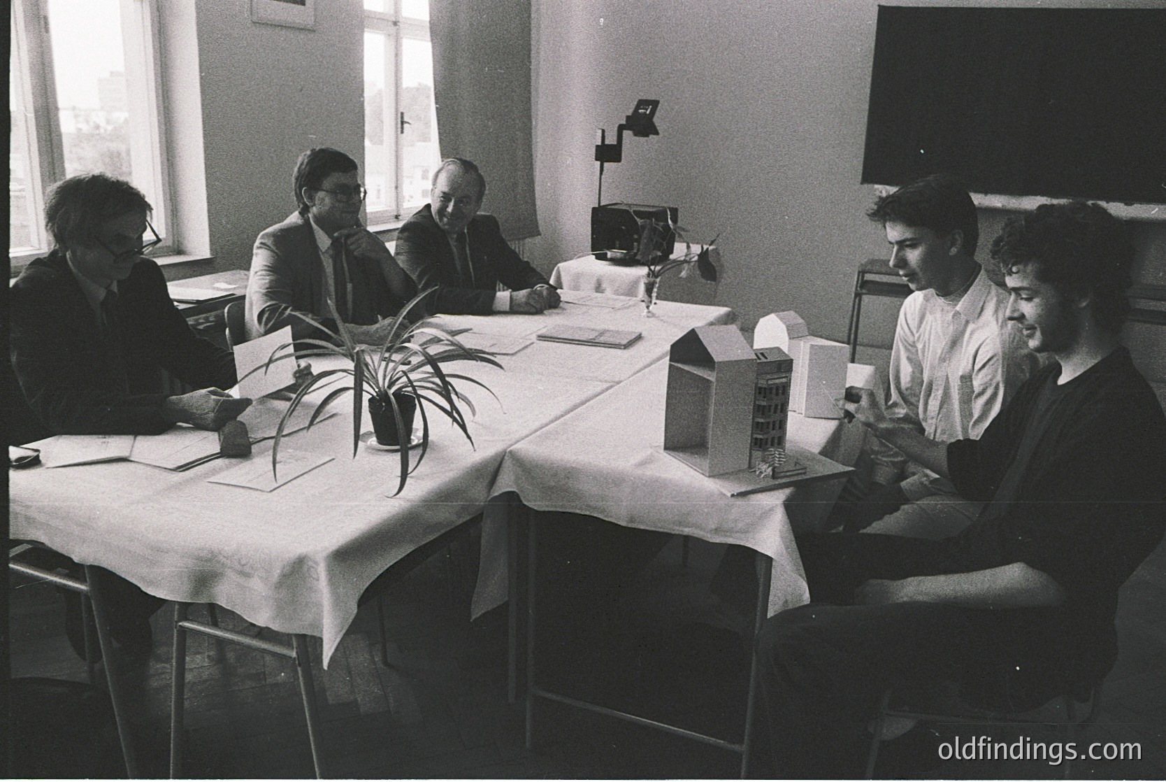 Mid-century conference room featuring five men in formal attire (suits, ties) reviewing architectural models and documents, 1960s–1970s. White tablecloths, potted plant, and overhead projector suggest a professional design discussion. Minimalist office decor reflects corporate or academic planning.