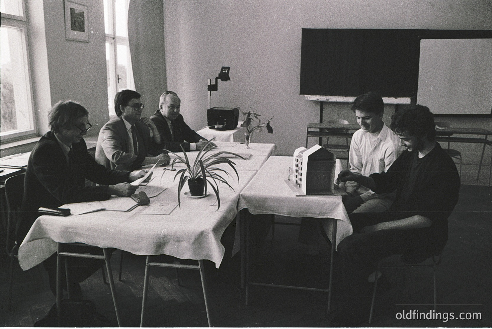 Four men in formal attire review architectural models and blueprints in a 1970s-era conference room, likely an East Bloc institution. Minimalist decor includes a potted plant, projector screen, and a small TV.