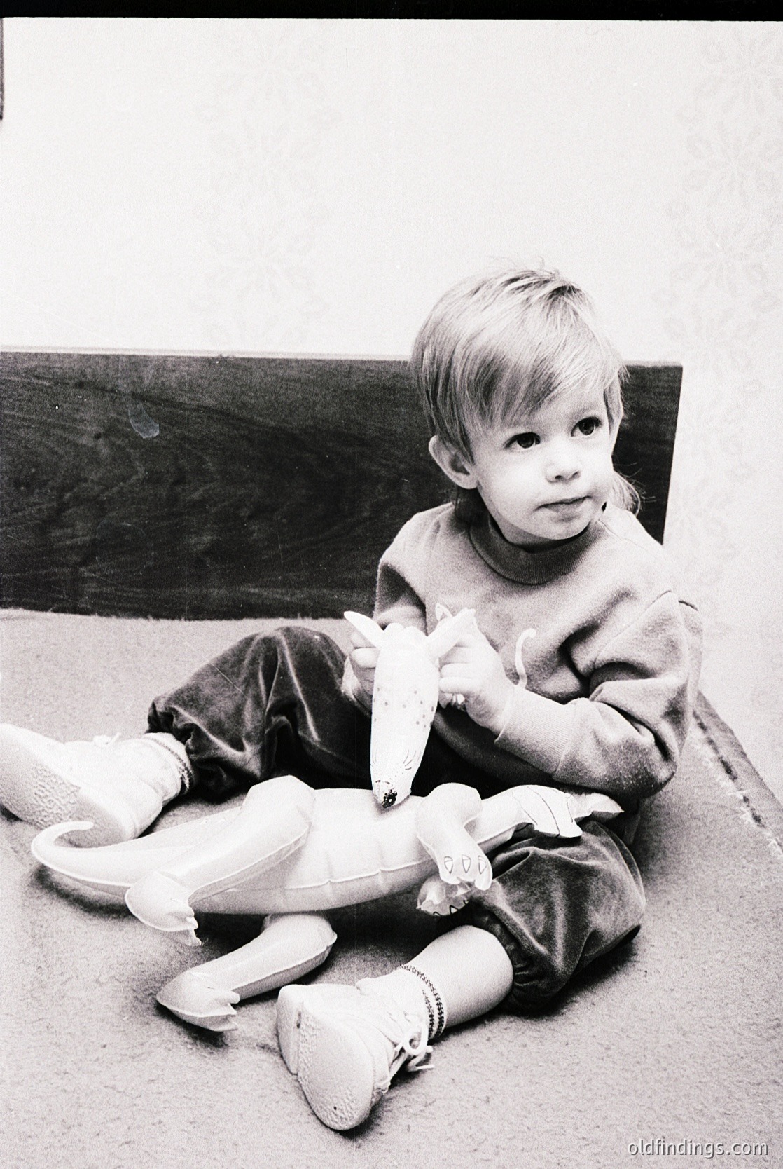 Vintage black-and-white photo of a young child (approx. 3-5 years) sitting on a carpeted floor, holding a stuffed duck toy. The child wears a sweater, pants, and white shoes with elastic bands. The setting appears domestic, likely mid-20th century (1950s-1960s).