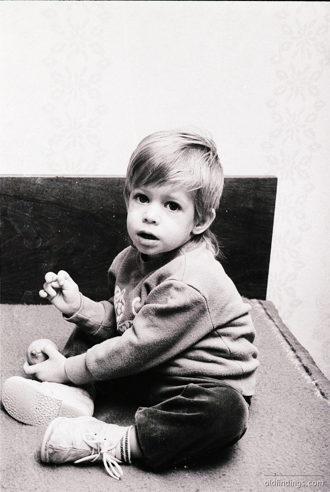 Young child in mid-20th-century clothing, sitting on a wooden floor with a wooden crate in background. Lighting suggests indoor domestic setting, likely 1950s–1960s.