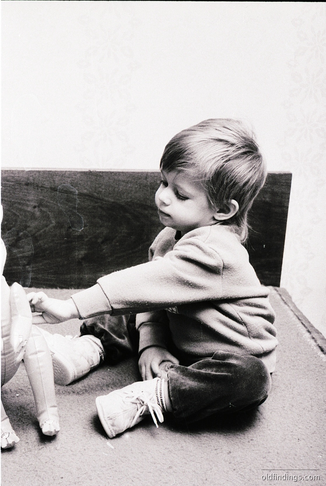 Young child in vintage sweater and knee-high socks plays with a doll on a wooden floor, likely mid-20th century. Classic black-and-white composition captures innocence and simplicity.