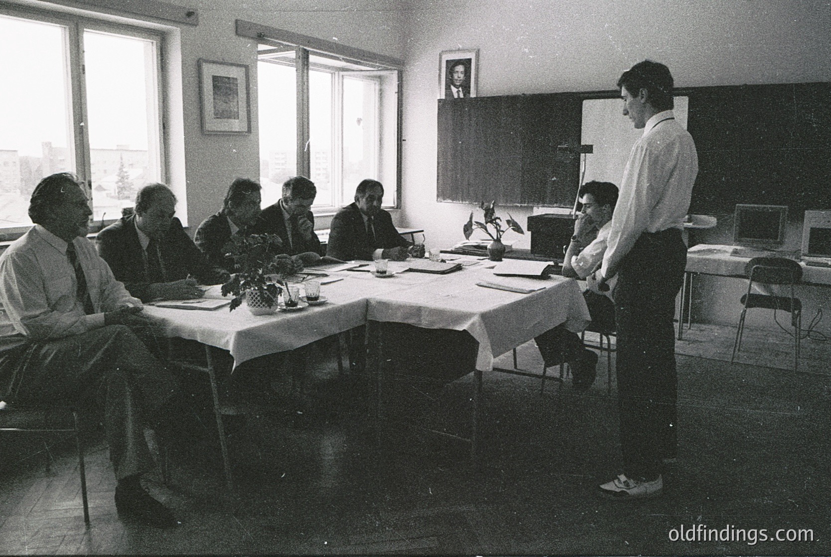 Mid-20th century office meeting in a functionalist-style room, likely Eastern Bloc era. Seven men in formal attire (suits, ties) seated around a rectangular table with white tablecloths. One man stands addressing the group; framed portrait on wall suggests institutional setting. Natural light floods through large windows.