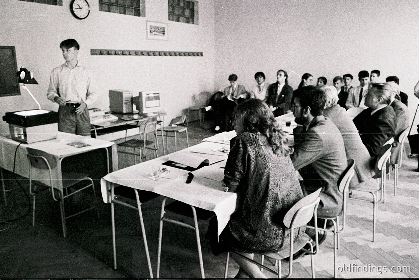 1970s classroom lecture hall with instructor at podium, students seated at long tables. Plain white walls, minimal decor, and utilitarian metal chairs. Likely Eastern Bloc-era educational setting.