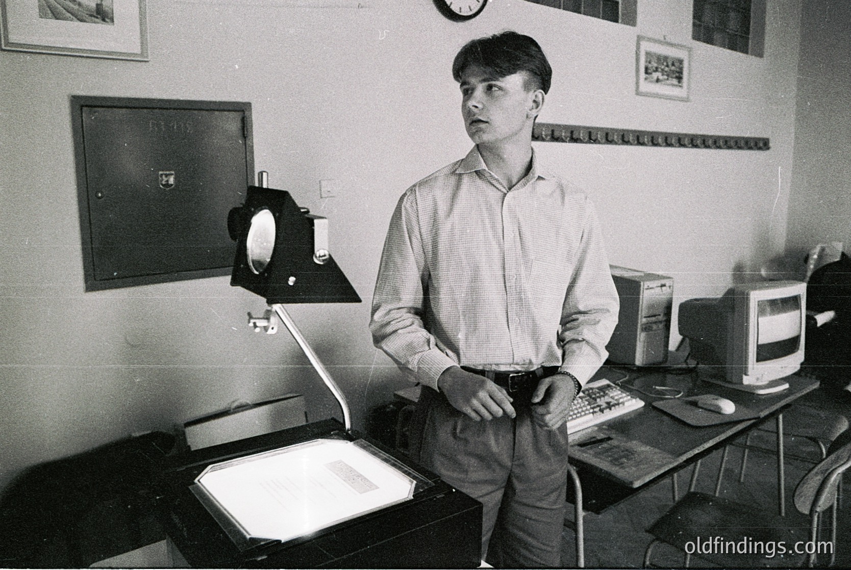 Man in a 1970s–80s office setting, standing beside a vintage slide projector and typewriter. White shirt, jeans, and a focused expression. Surrounding equipment includes a CRT monitor, tape measure, and framed photos on walls. Industrial, retro workspace aesthetic.