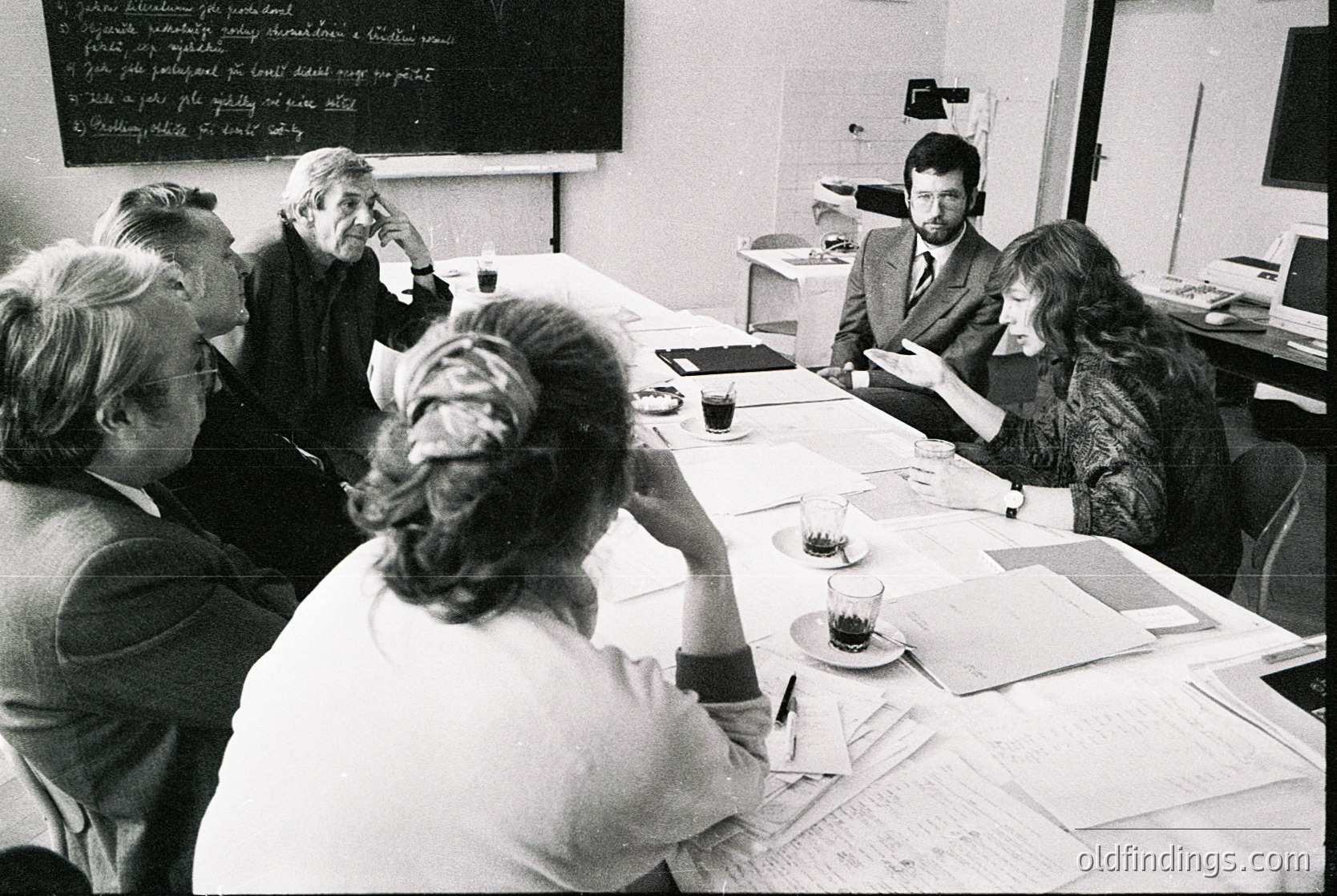 A 1970s-era office meeting featuring six professionals in formal attire around a rectangular table. Black-and-white chalkboard and papers suggest academic or corporate discussion. Glass of tea and pens indicate a collaborative session. Minimalist office decor with wooden paneling.
