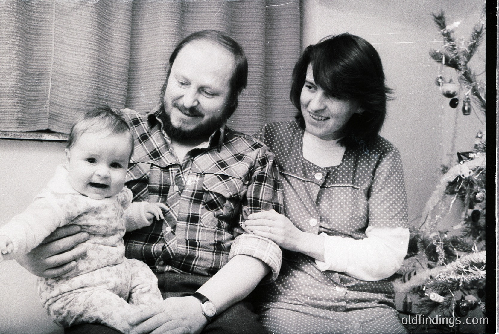 A black-and-white family portrait from the 1970s–1980s, featuring a man in a plaid shirt, a woman in a patterned blouse, and a baby in a light sweater. They pose indoors near a decorated Christmas tree with ornaments. Warm, candid expressions capture holiday joy.