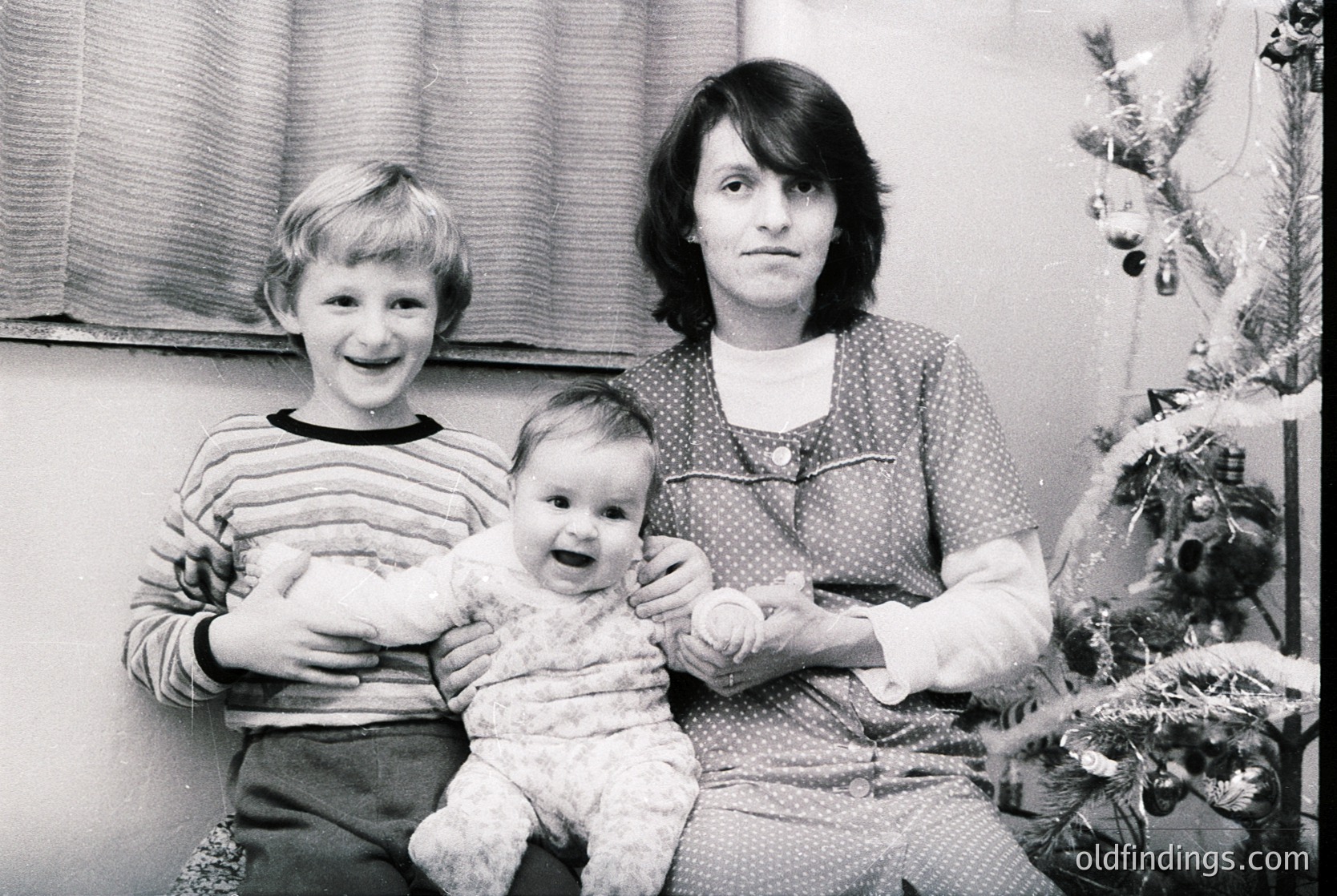 Family portrait featuring a woman holding a baby, with a young boy standing beside her. Indoor setting with a decorated Christmas tree in the background, suggesting late 20th-century holiday traditions. Clothing styles and black-and-white tone indicate **1970s-1980s**. [Family holiday portrait, 1970s-1980s, Christmas tree, mother and child, black & white photography]