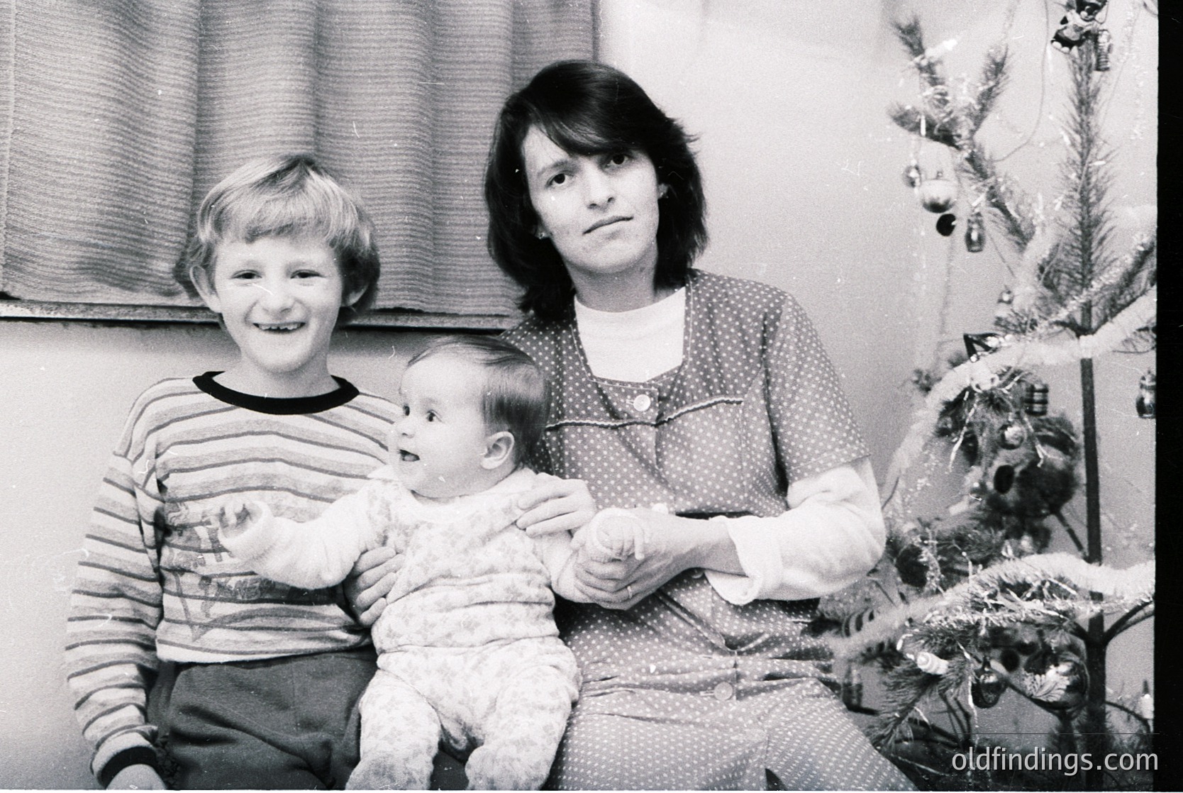 A 1970s family portrait featuring a woman holding a baby, with a smiling older child beside her. Indoor setting with a decorated Christmas tree in the background. Warm, candid expressions capture holiday warmth.