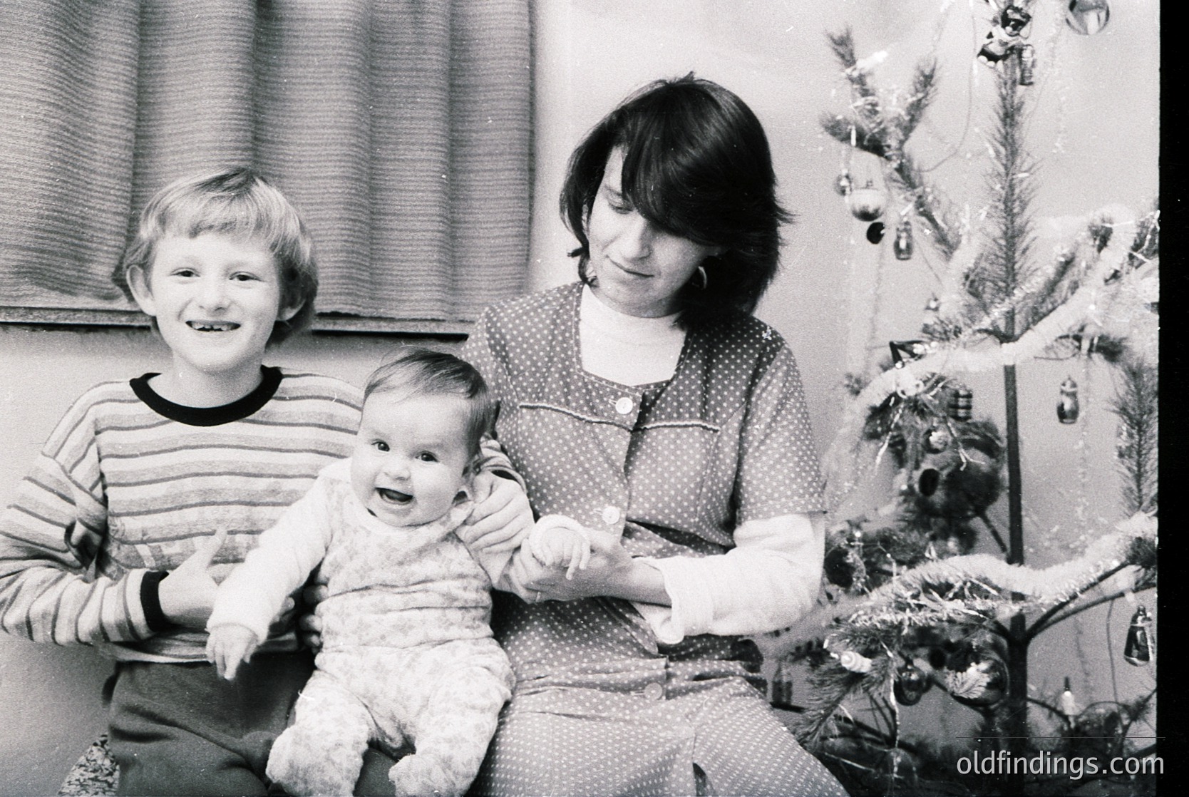 Family portrait featuring a woman holding a baby, with a young boy standing beside them. Decorated Christmas tree with ornaments and lights in the background. Mid-20th century (likely 1960s–1970s) holiday interior.