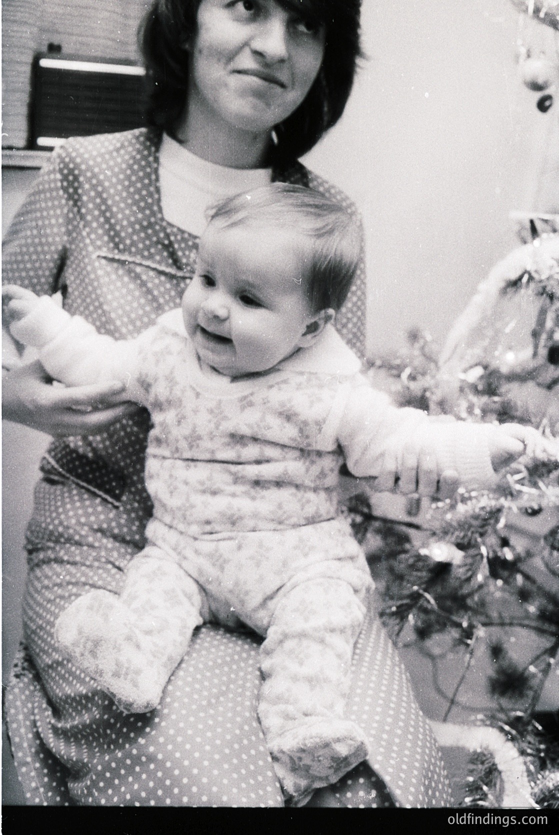A woman in a polka-dot blouse holds a toddler in a floral onesie, both posing indoors near a decorated Christmas tree. The scene reflects mid-20th-century holiday traditions, likely 1950s–1960s.