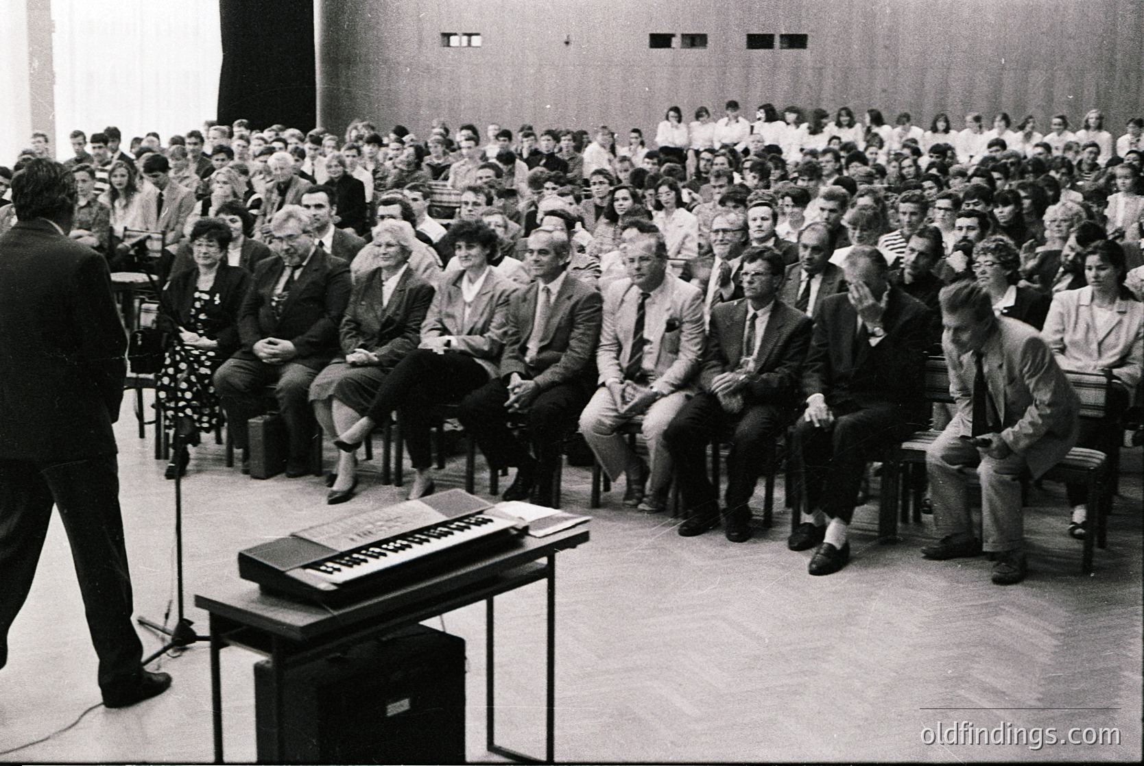 Black-and-white lecture hall scene with speaker addressing seated audience of ~150 individuals in formal attire (1960s–70s). Wooden podium holds sheet music and microphone; tiered seating arranged in rows. Minimalist interior with plain walls and carpeted floor.