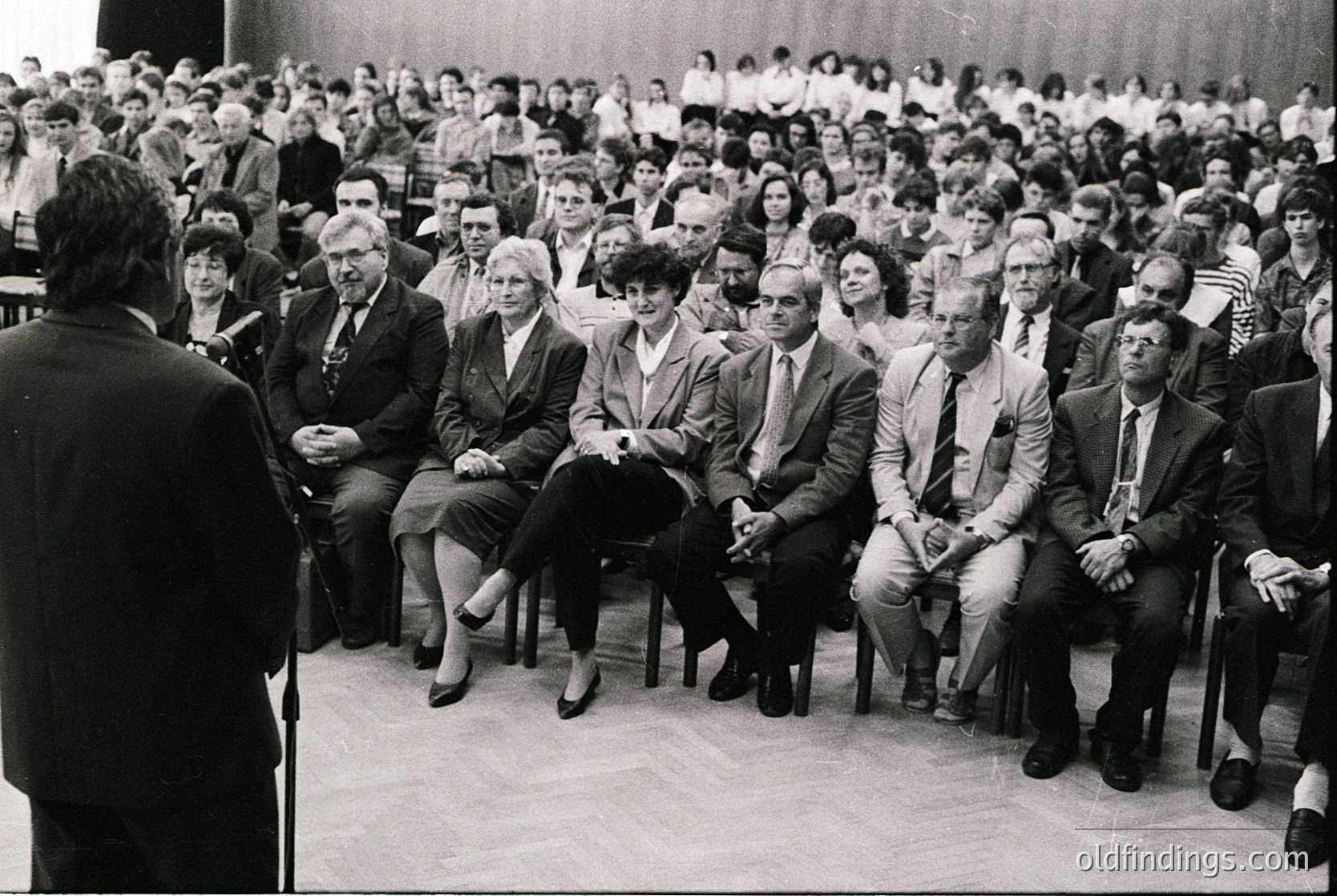 Black-and-white photo of a formal indoor lecture or conference, likely mid-20th century. Audience seated in tiered rows, men in suits and women in dresses, attentive to a speaker at a podium. Architectural details include wooden chairs and a neutral-toned stage area. Crowd density suggests institutional or governmental event.